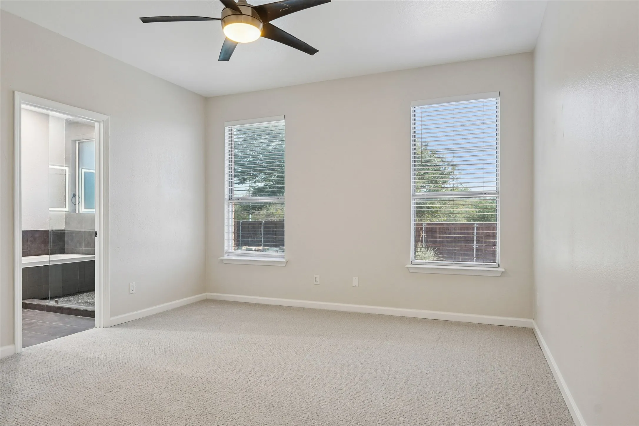 Carpeted flooring, two windows with blinds, modern ceiling fan with integrated lighting, and an attached full bathroom with a tiled tub