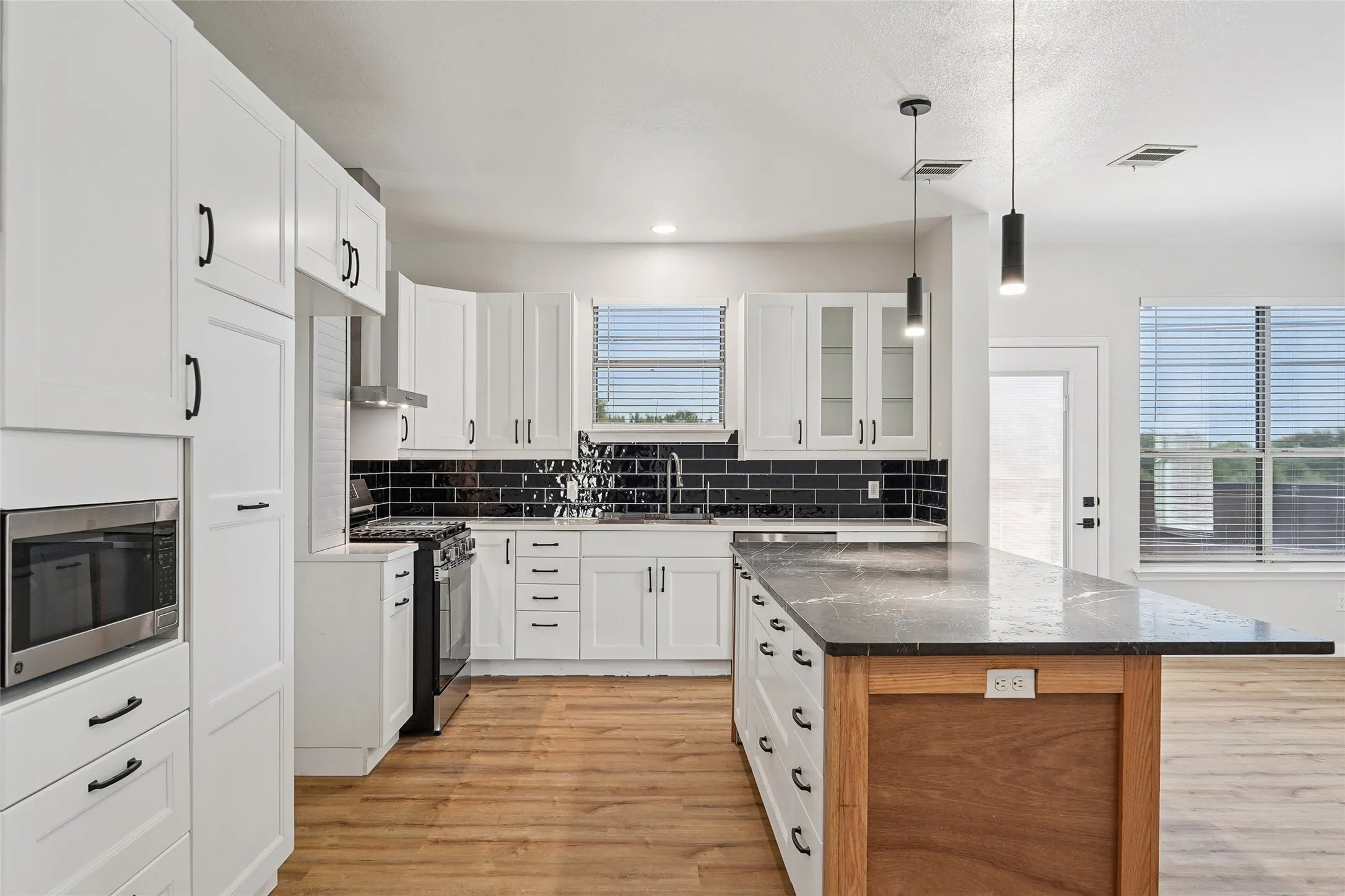 White cabinetry, black subway tile backsplash, stainless steel appliances, wood-finish flooring, and a kitchen island with pendant lighting