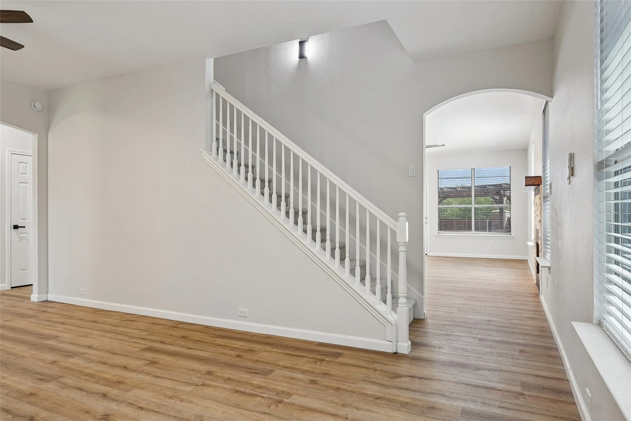 Wood-finish flooring, white painted walls, white spindle staircase, arched doorway, and a window with blinds