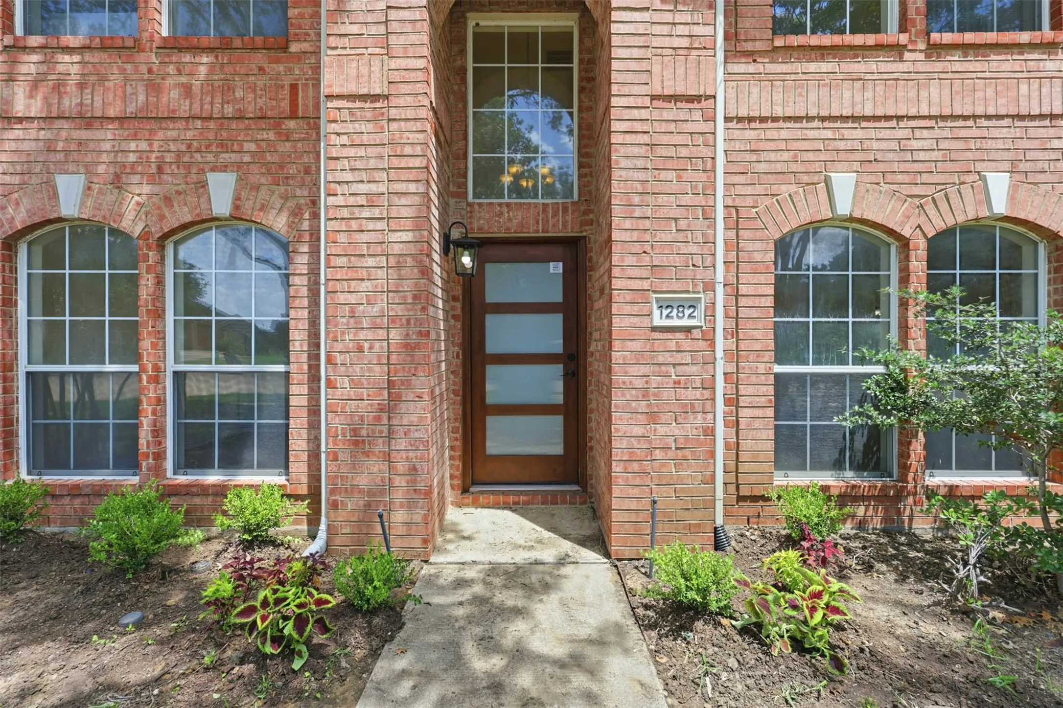 Brick facade featuring a recessed entry, arched windows with gridded panes, a contemporary wood-finish door with frosted glass panels, and exterior lighting fixtures