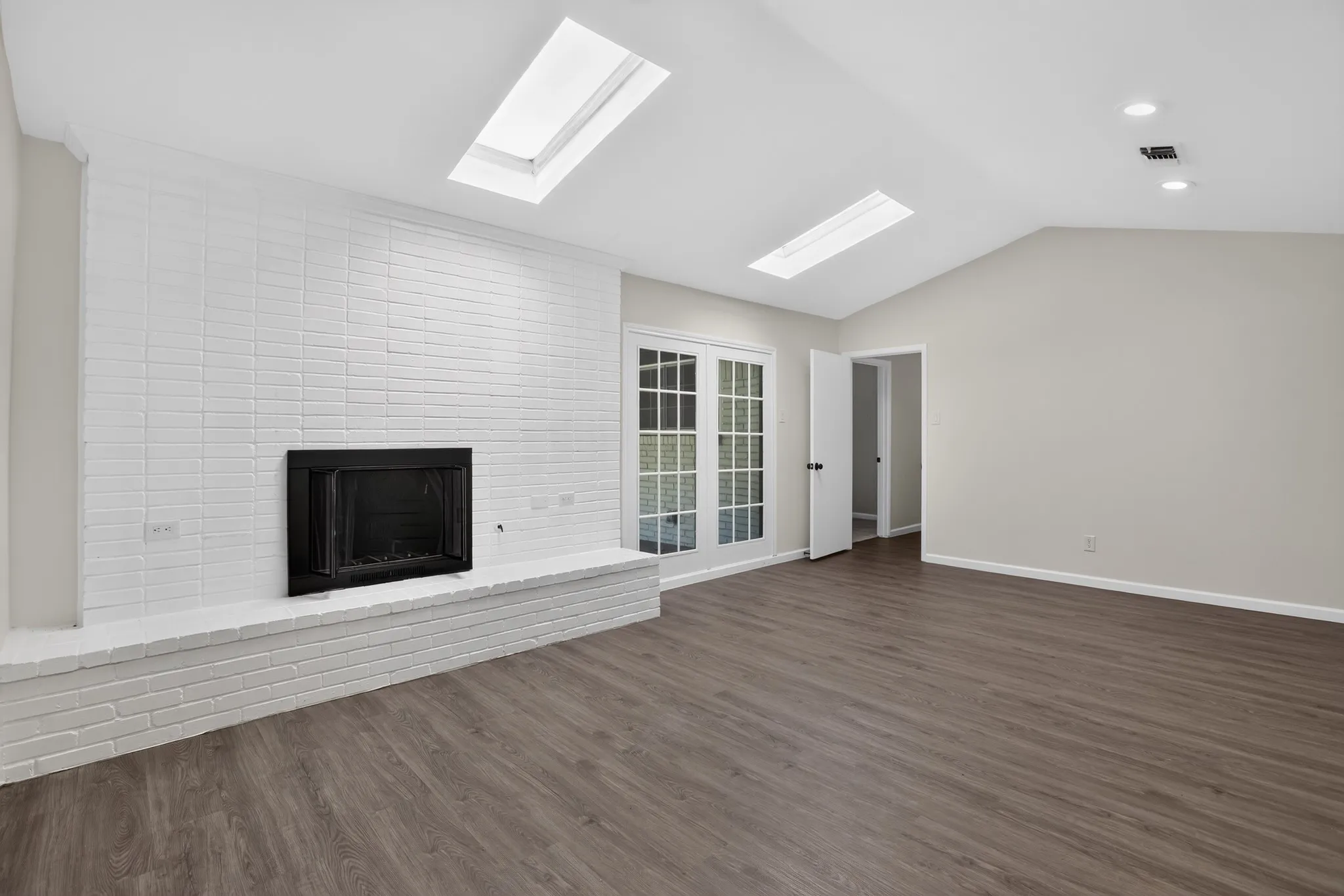 Spacious living area featuring a white brick fireplace, vaulted ceiling, two skylights, and wood-finish flooring