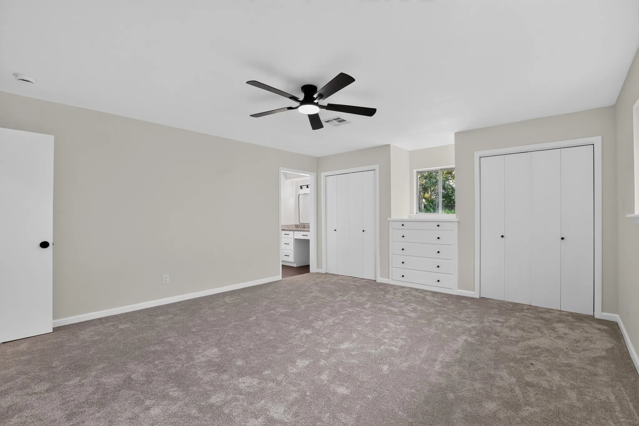 Spacious carpeted room featuring a ceiling fan with integrated lighting, multiple white bi-fold closet doors, a built-in five-drawer dresser, and a window
