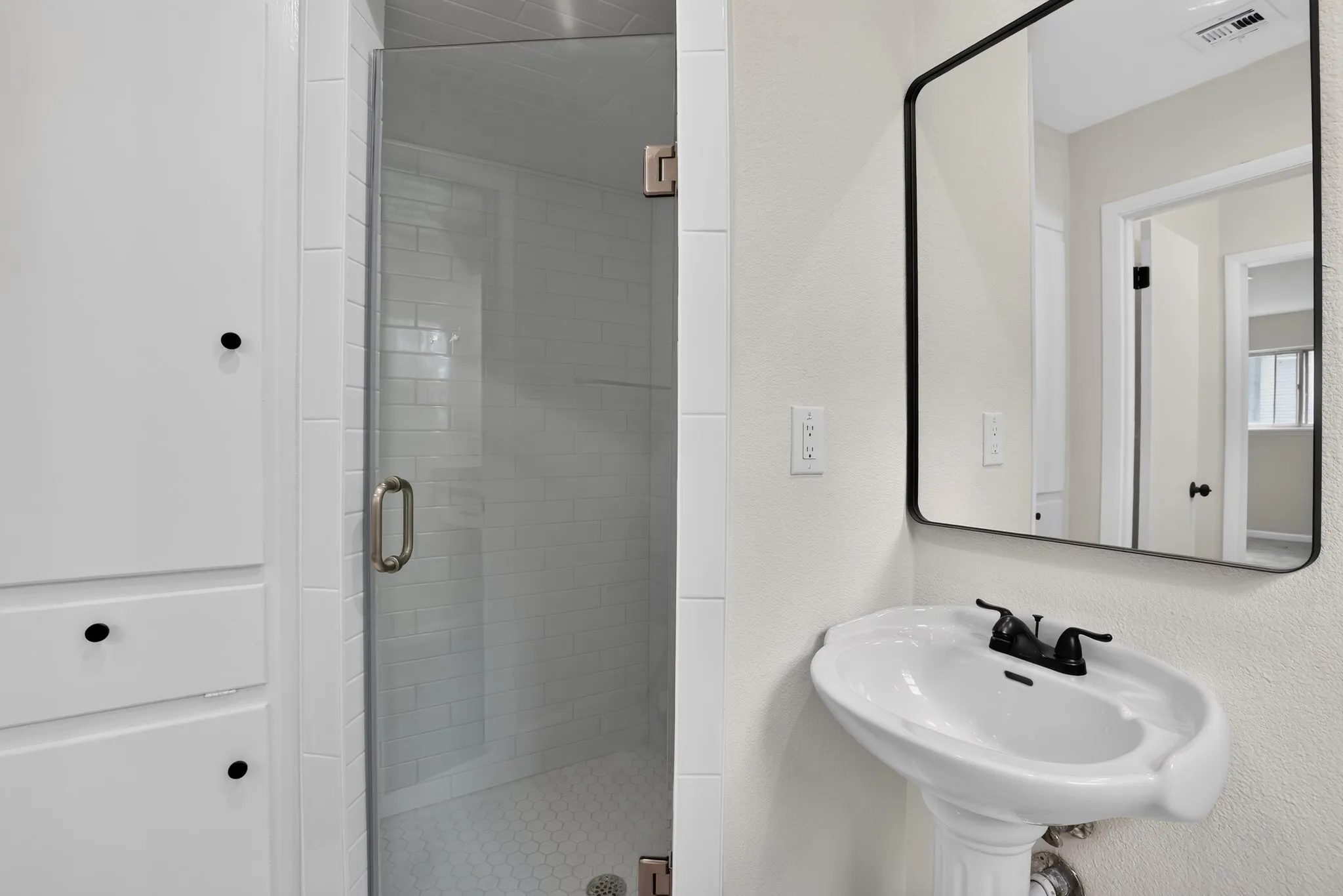 Bathroom featuring a clear glass shower enclosure, white subway tile shower surround, hexagonal tile shower floor, and a pedestal sink with a black-framed mirror