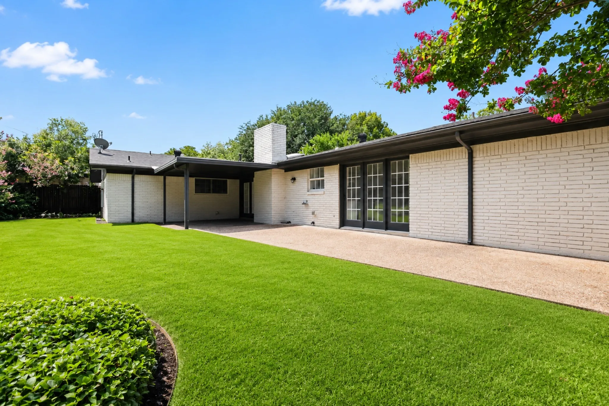 Expansive backyard featuring a manicured lawn, an exposed aggregate patio, white brick exterior, black-framed French doors, and a covered patio area