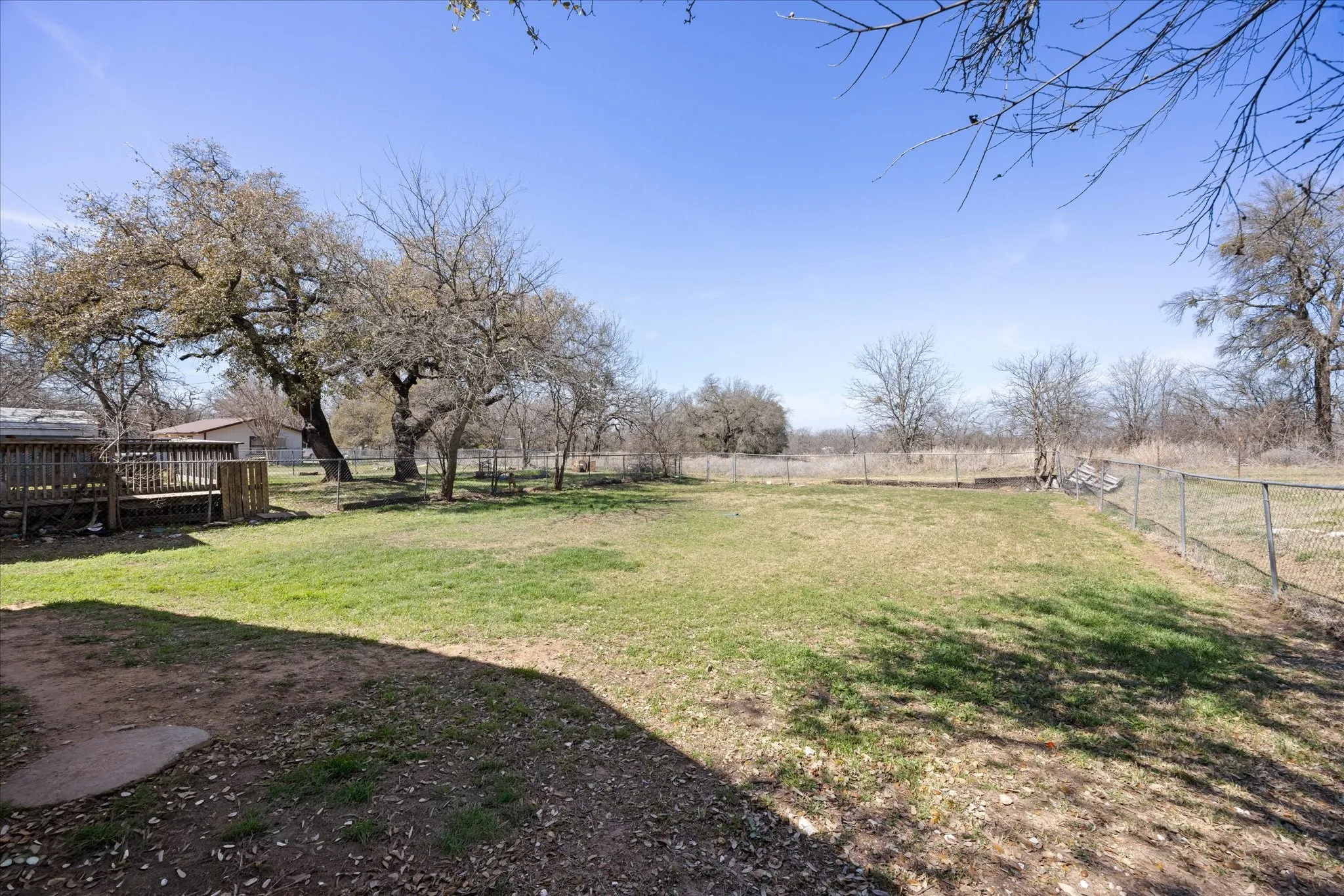 Expansive outdoor area featuring a wooden deck, mature trees, a chain-link perimeter fence, and a grassy lawn