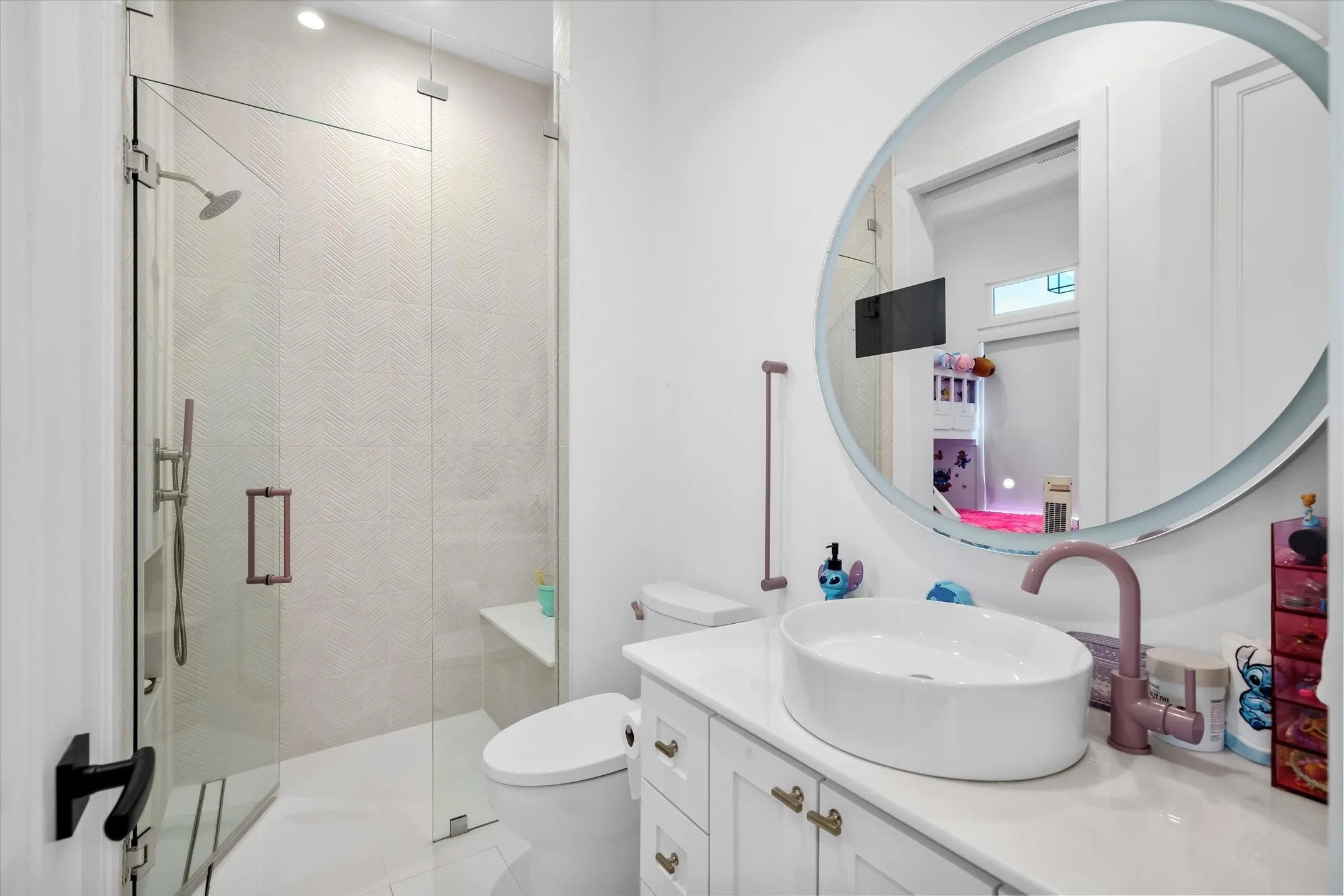 Bathroom featuring a frameless glass shower enclosure with a built-in bench, chevron-patterned shower wall tiles, a round illuminated mirror, a vessel sink, and a white vanity with gold-tone hardware