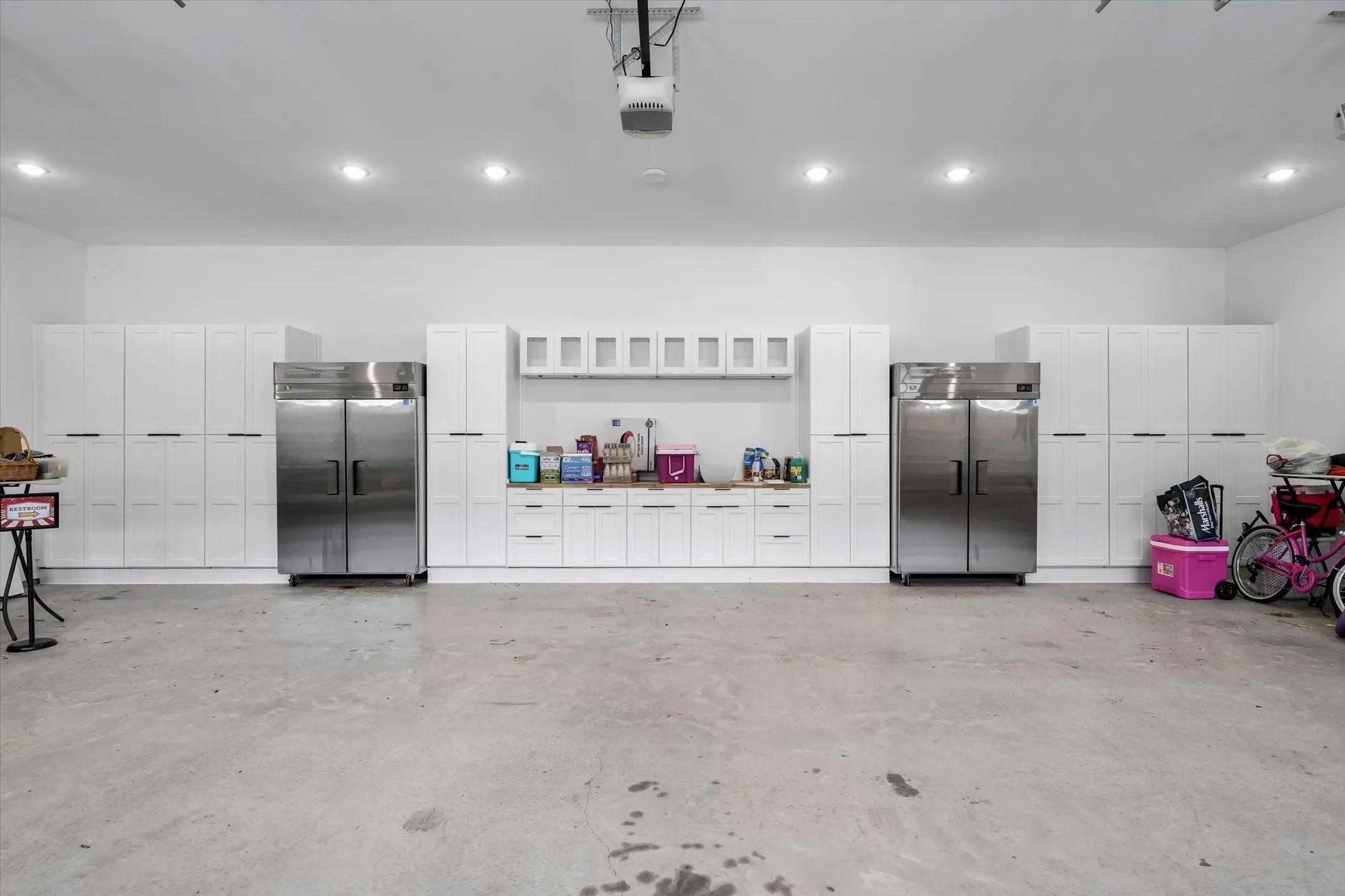 Spacious garage featuring extensive built-in white cabinetry, two large stainless steel refrigerators, and recessed ceiling lighting