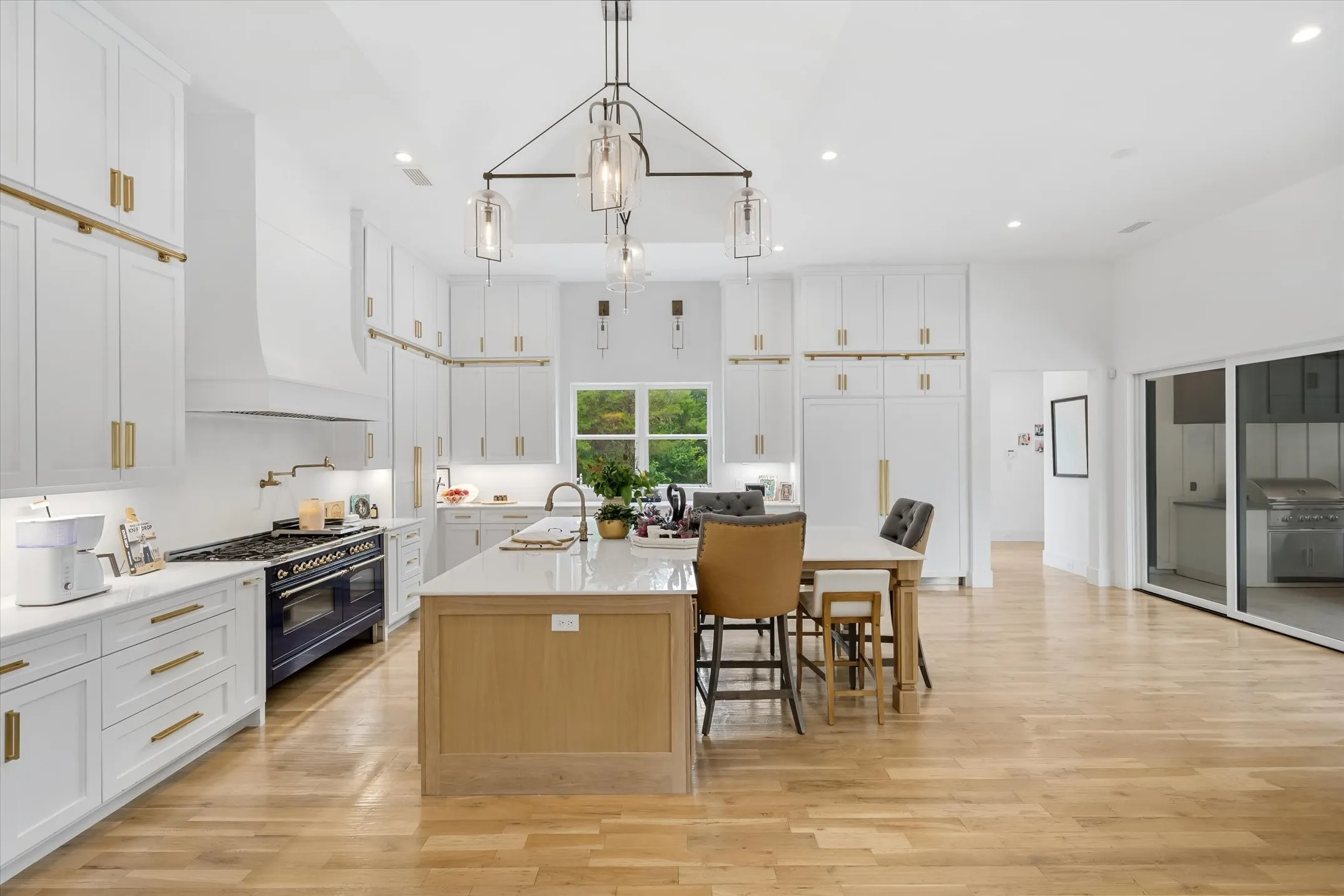 Gourmet kitchen featuring a substantial central island, professional-grade range, abundant white cabinetry with gold-tone hardware, wood-finish flooring, and a vaulted ceiling with a distinctive linear chandelier