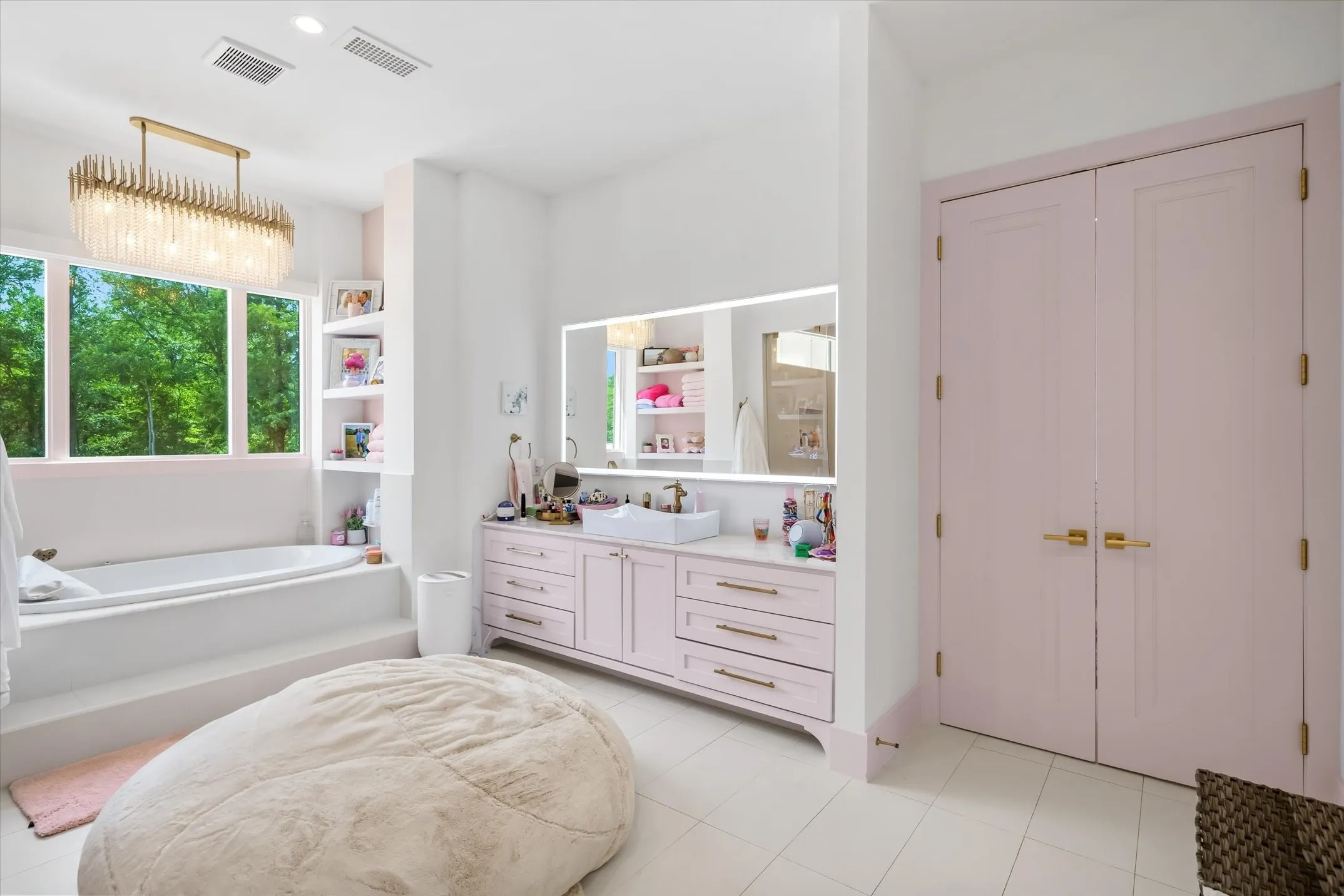 Bright bathroom featuring a large soaking tub with a stepped platform, an expansive vanity with pink cabinetry and gold-tone hardware, a rectangular vessel sink, and a large backlit mirror