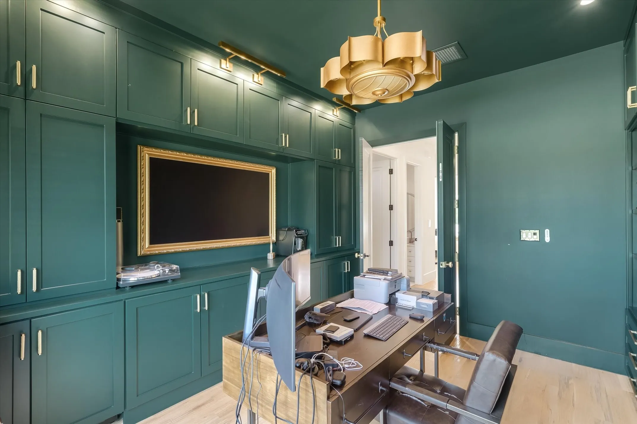 Built-in cabinetry in a deep green finish with gold hardware, integrated media niche, gold-toned ceiling fixture, light wood-finish flooring, and a solid wood desk, Murphy bed