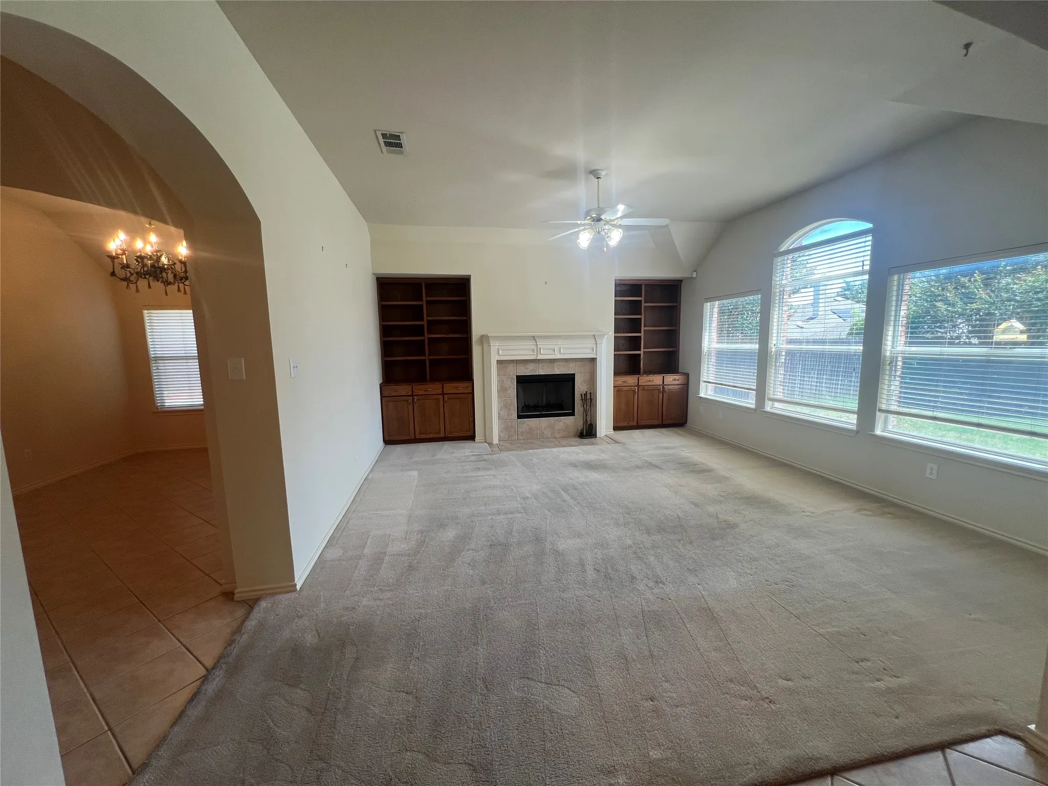 Vaulted ceiling with ceiling fan, large arched windows, fireplace with mantel, built-in shelving, and carpet flooring