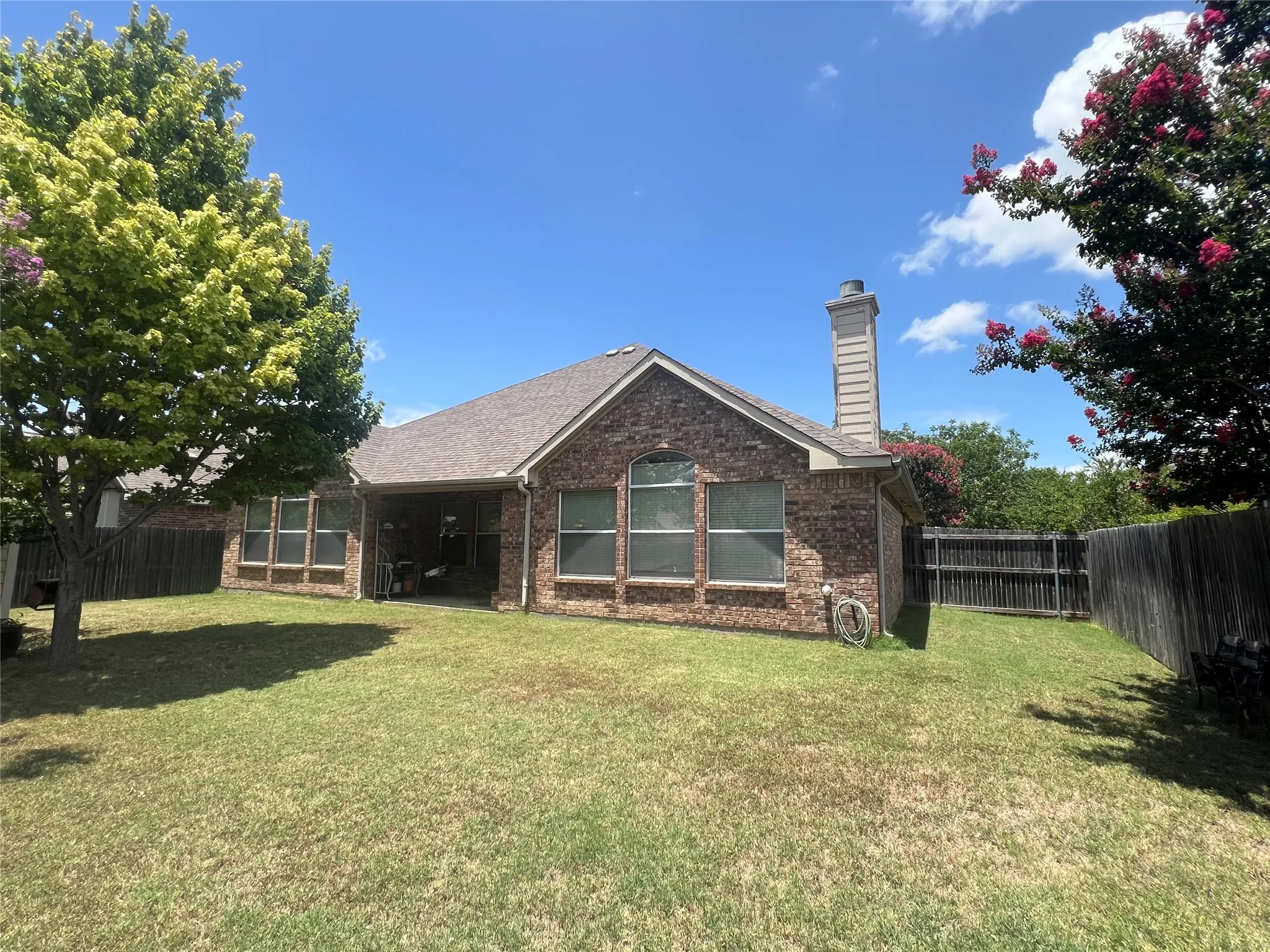 Expansive backyard featuring a covered patio, mature tree, brick exterior, multiple windows, and privacy fencing