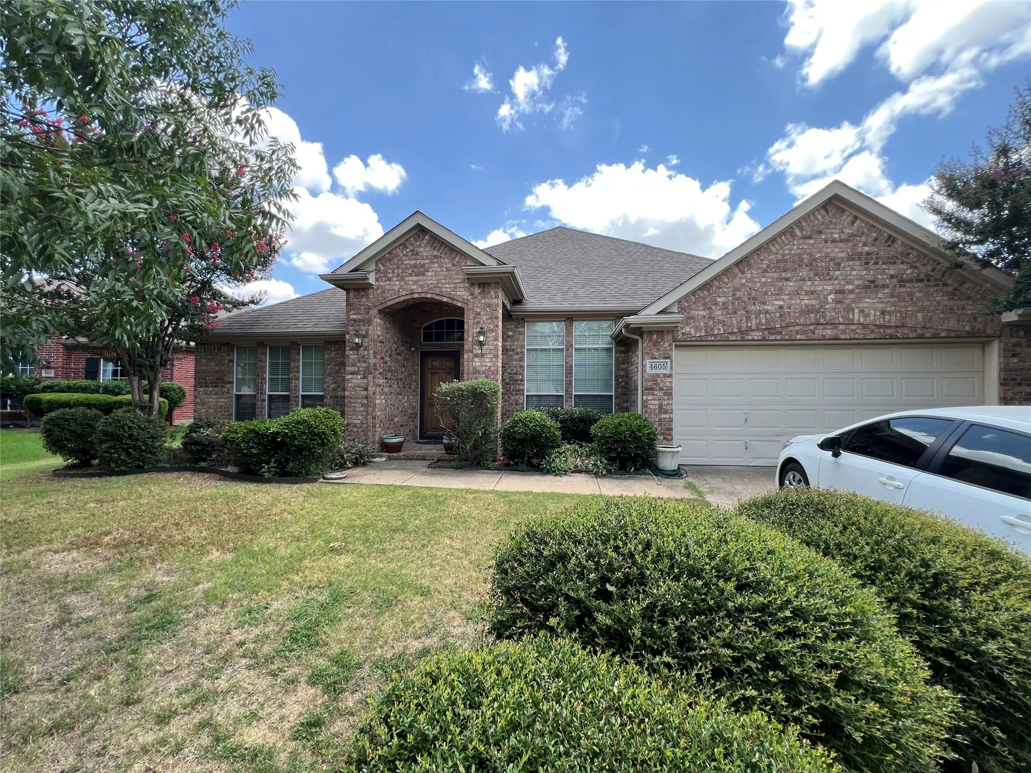 Brick facade residence featuring an arched entryway, gabled roofline, front-facing windows, attached garage, and mature landscaping