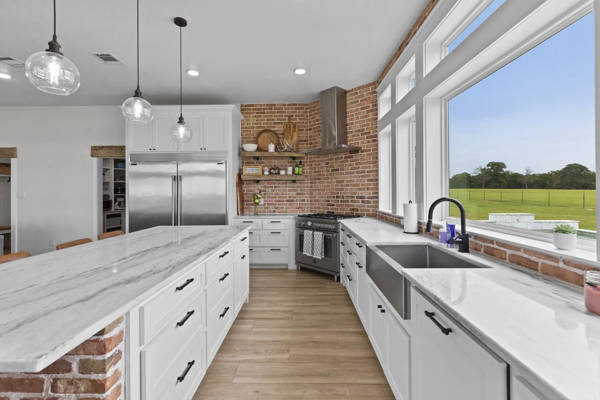 Kitchen featuring an expansive center island with a brick base and stone countertop, wood-finish flooring, white cabinetry with black hardware, stainless steel appliances, and a brick accent wall