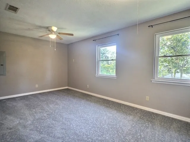 Spacious room featuring neutral painted walls, two windows with white trim, a ceiling fan with integrated lighting, and gray patterned carpeting