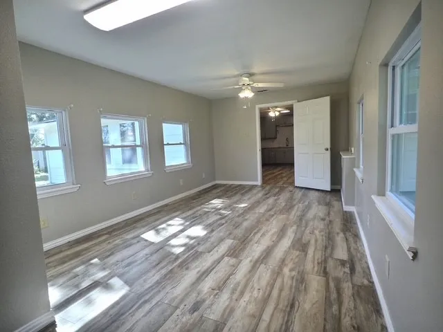 Spacious room featuring wood-finish flooring, light grey painted walls, multiple white-trimmed windows, and a ceiling fan with integrated lighting