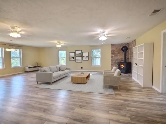 Spacious living area featuring wood-finish flooring, multiple windows, a brick accent wall with a wood-burning stove, and ceiling fans