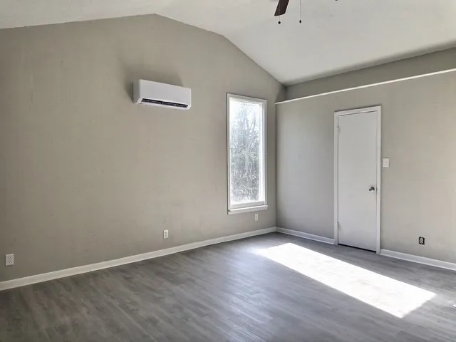 Spacious room featuring vaulted ceilings, wood-finish flooring, a wall-mounted mini-split AC unit, and a ceiling fan