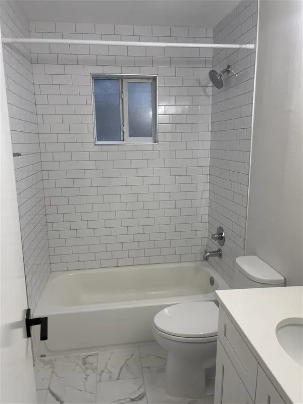 Bathroom featuring a white subway tile shower surround, a built-in window with frosted panes, chrome shower fixtures, a white bathtub, and marble-finish tile flooring