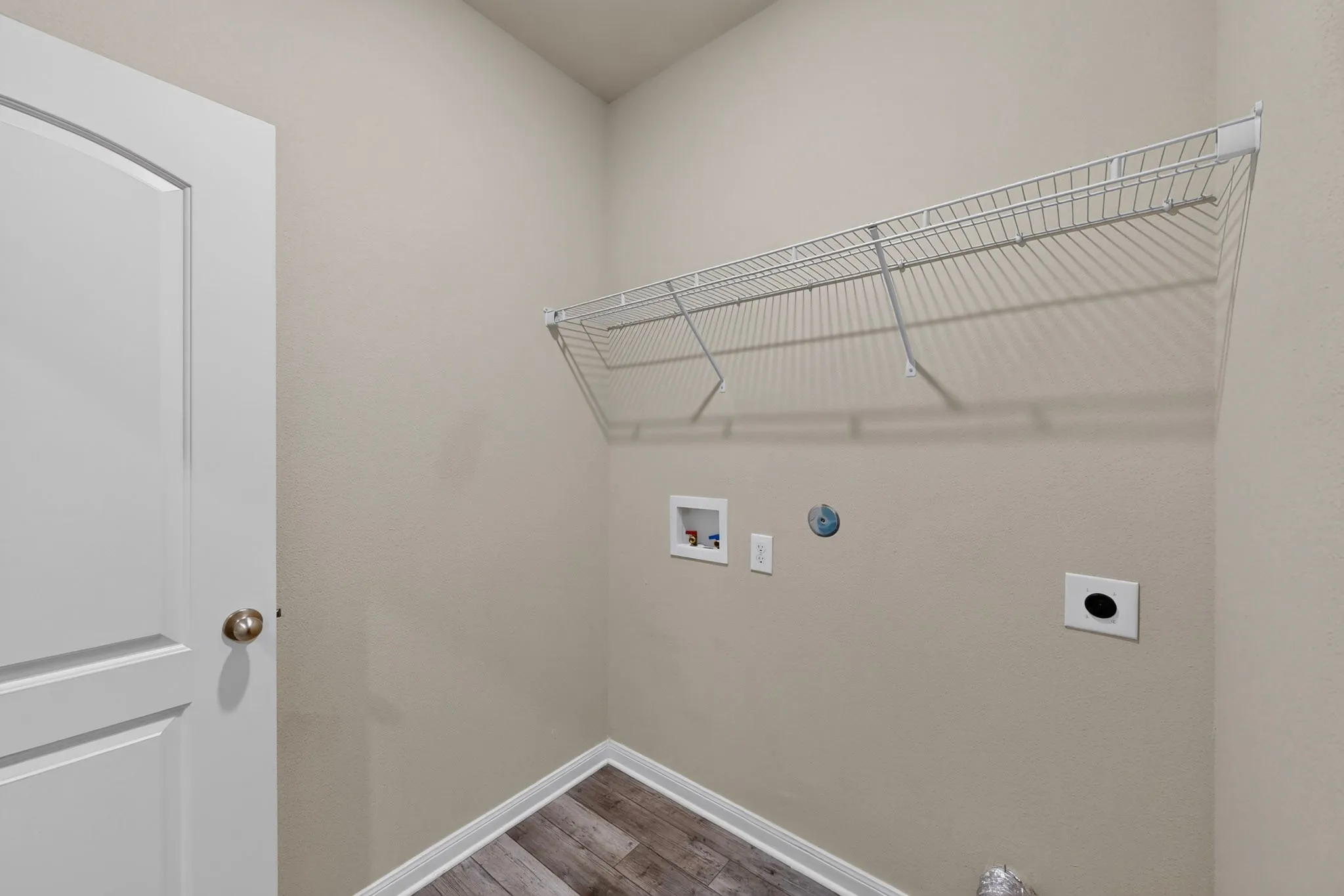 Laundry area featuring wood-finish flooring, light neutral wall paint, a white wire shelving unit, and a white paneled door with an arched top trim