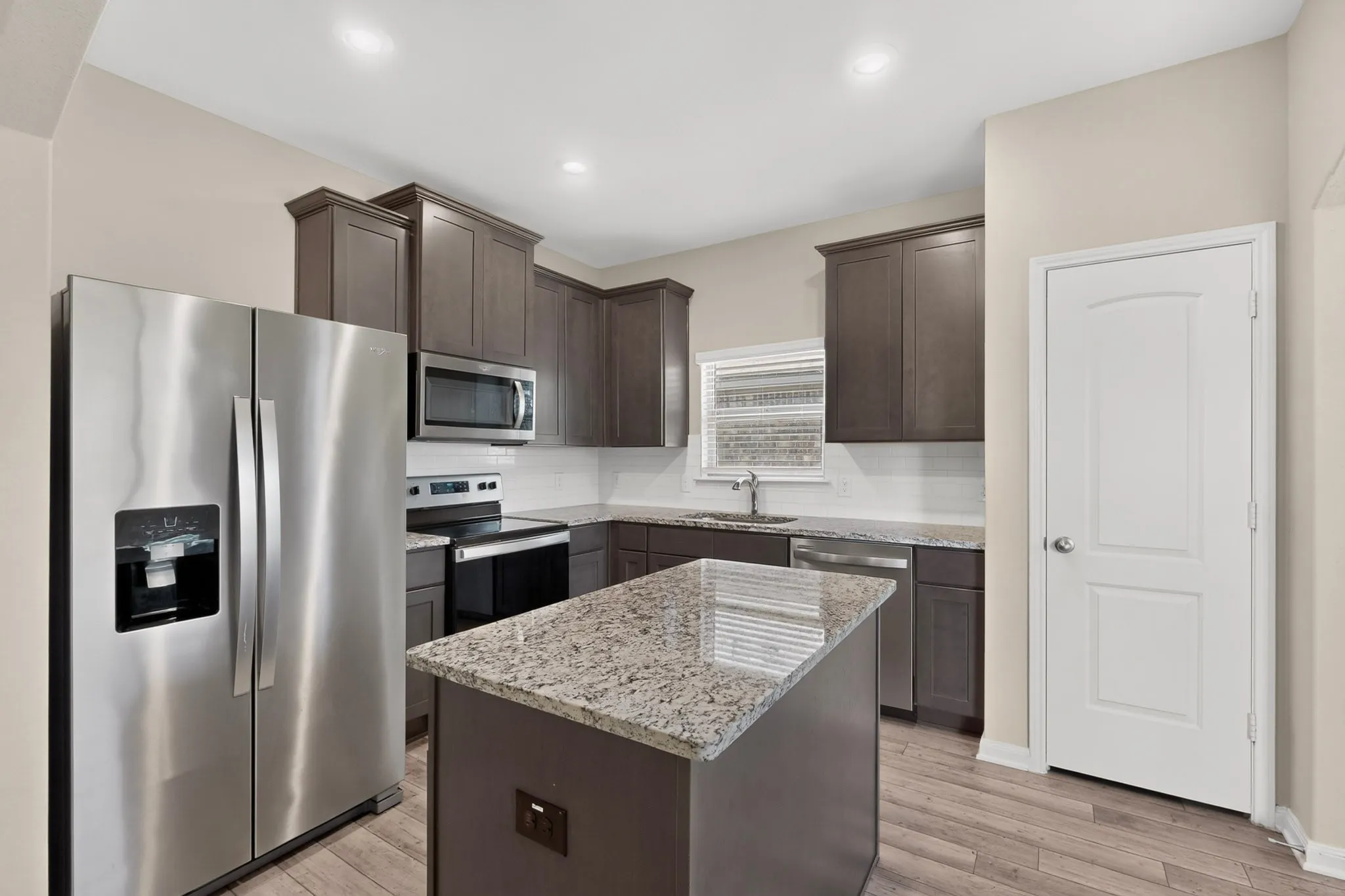 Contemporary kitchen featuring an island with a granite countertop, stainless steel appliances, dark wood-finish cabinetry, subway tile backsplash, and wood-finish flooring