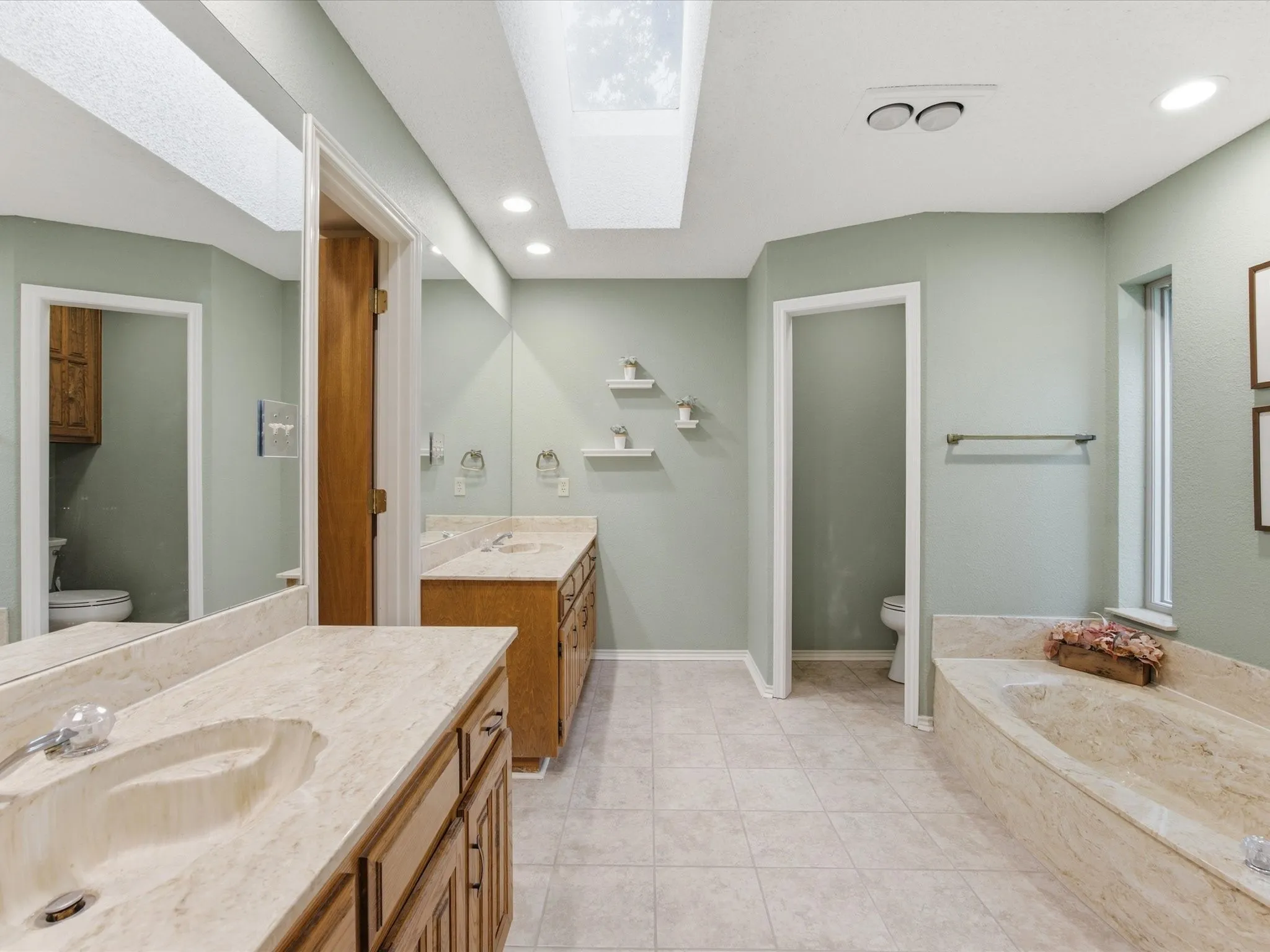 Primary bathroom featuring a large skylight, dual vanity with integrated sinks, wood-finish cabinetry, a built-in tub with stone-finish surround, and tiled flooring