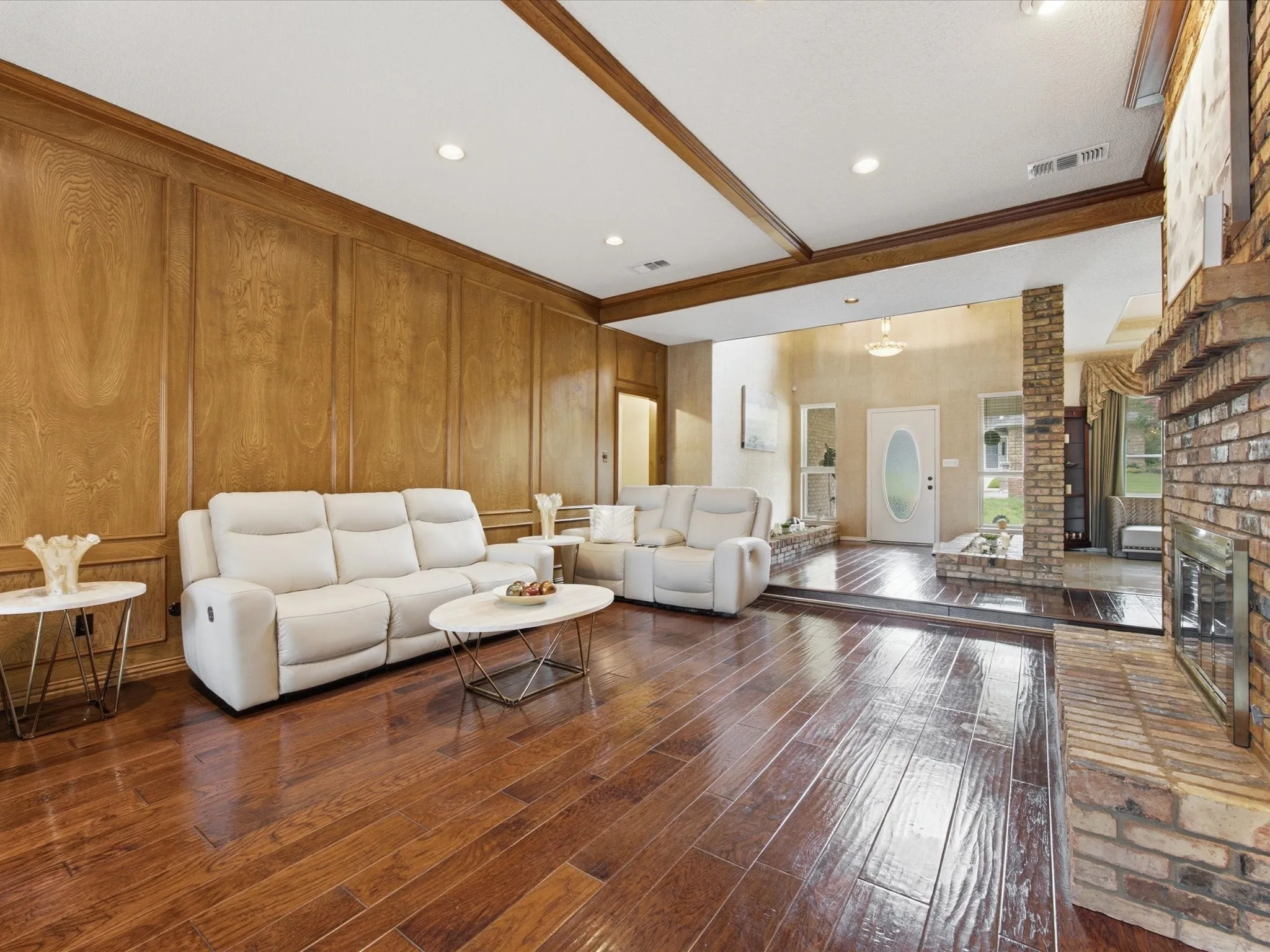 Spacious living area featuring wood-finish flooring, wood paneling, a brick fireplace, exposed wood ceiling beams, and recessed lighting