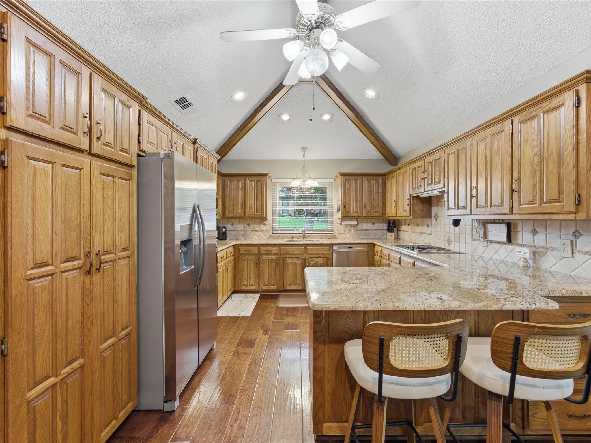Kitchen featuring a vaulted ceiling with exposed wood beams, extensive wood cabinetry, wood-finish flooring, a stainless steel refrigerator, and stone countertops