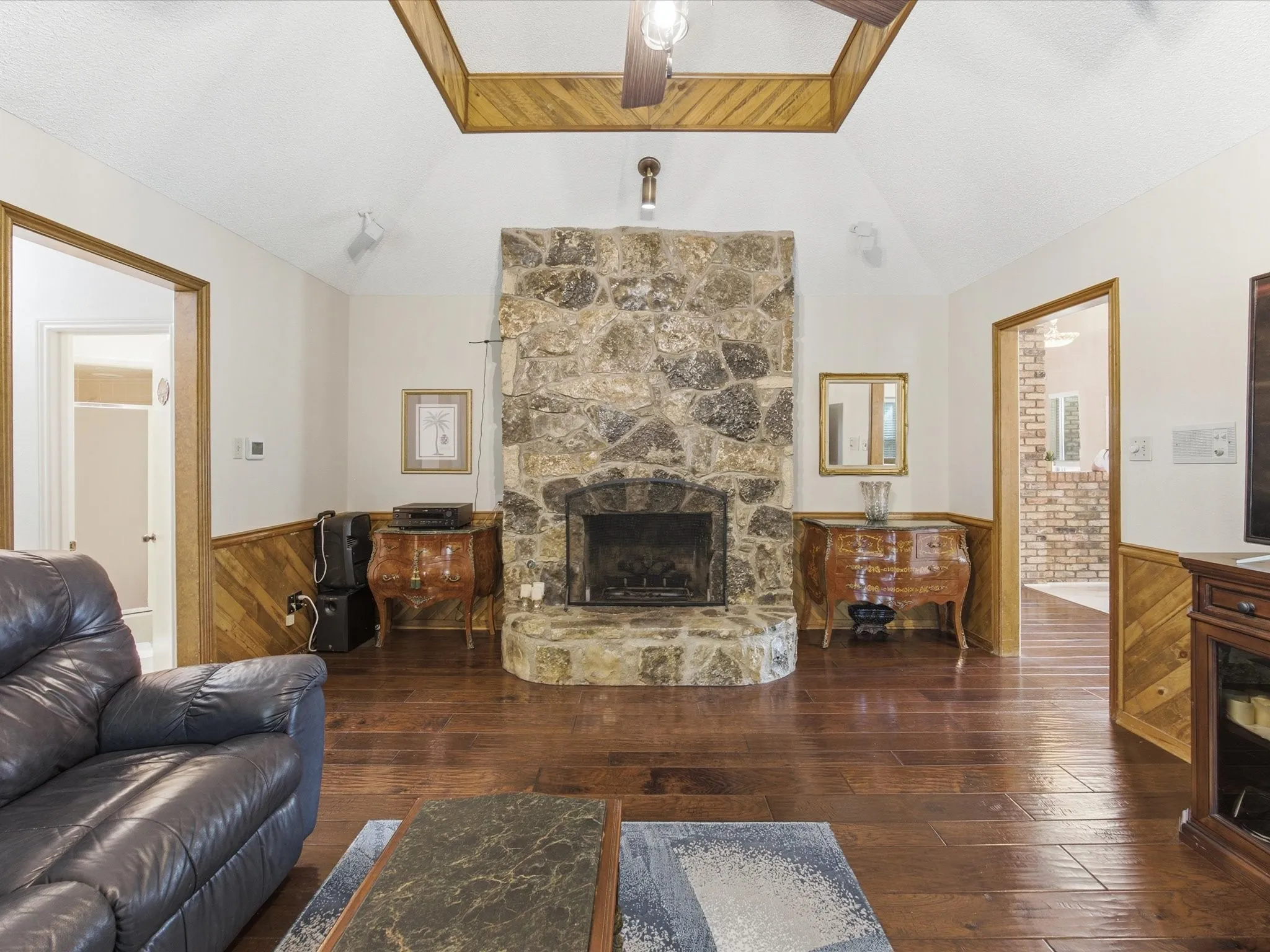 Spacious living area featuring a stone fireplace with hearth, vaulted ceiling, wood-finish flooring, and wood paneling on lower walls