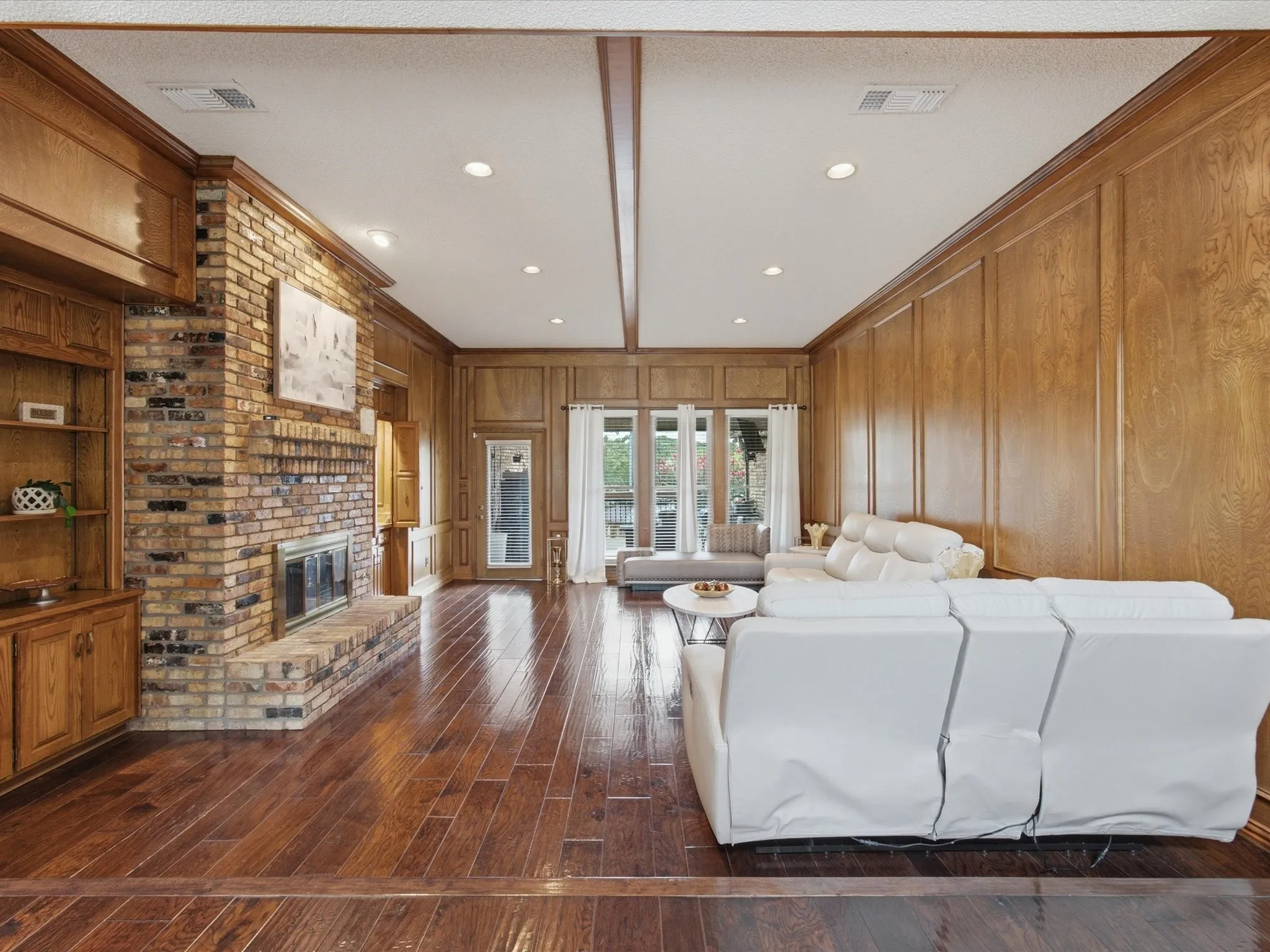 Spacious living area featuring extensive wood paneling, a brick fireplace, built-in shelving, wood-finish flooring, and recessed lighting