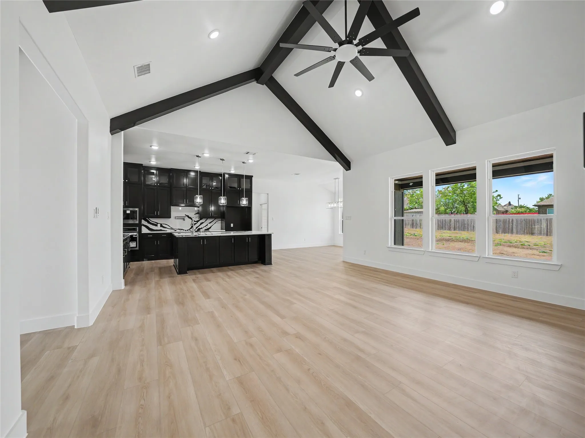 Vaulted ceiling with exposed black beams and ceiling fan, light wood-finish flooring, and a kitchen featuring black cabinetry, a white and black veined countertop, and pendant lighting