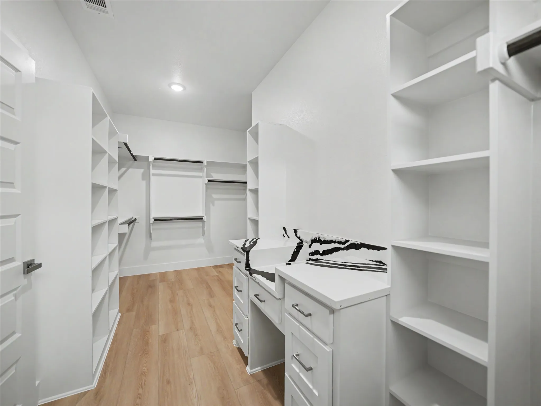 Spacious walk-in closet featuring light wood-finish flooring, recessed lighting, extensive white shelving, multiple clothing rods, and a built-in white vanity with drawers
