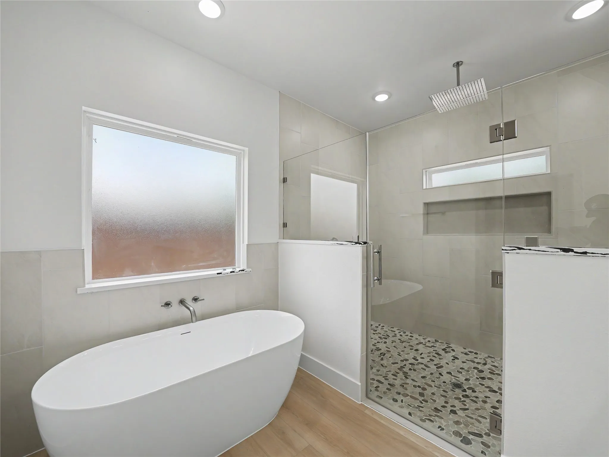 Freestanding soaking tub with wall-mounted faucet, alongside a glass-enclosed shower featuring a rainfall showerhead, pebble-tiled floor, and recessed shelf