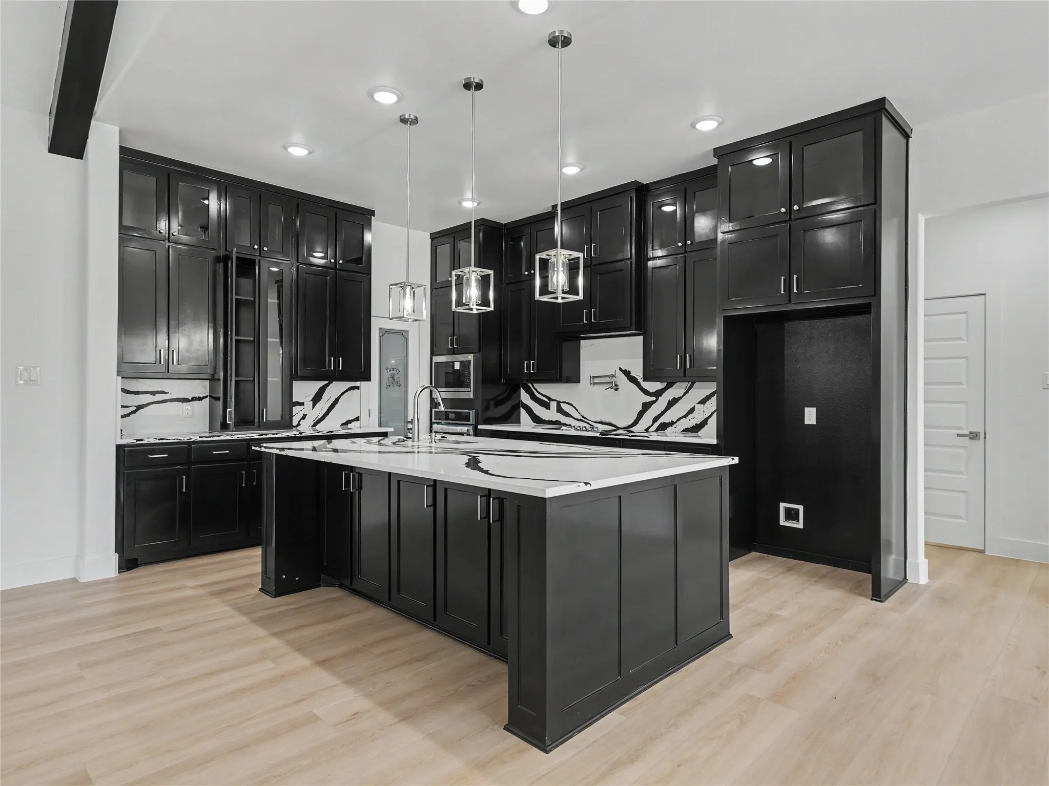 Gourmet kitchen featuring a large central island with integrated sink, black cabinetry with glass-front uppers, white countertops, and a striking black and white backsplash