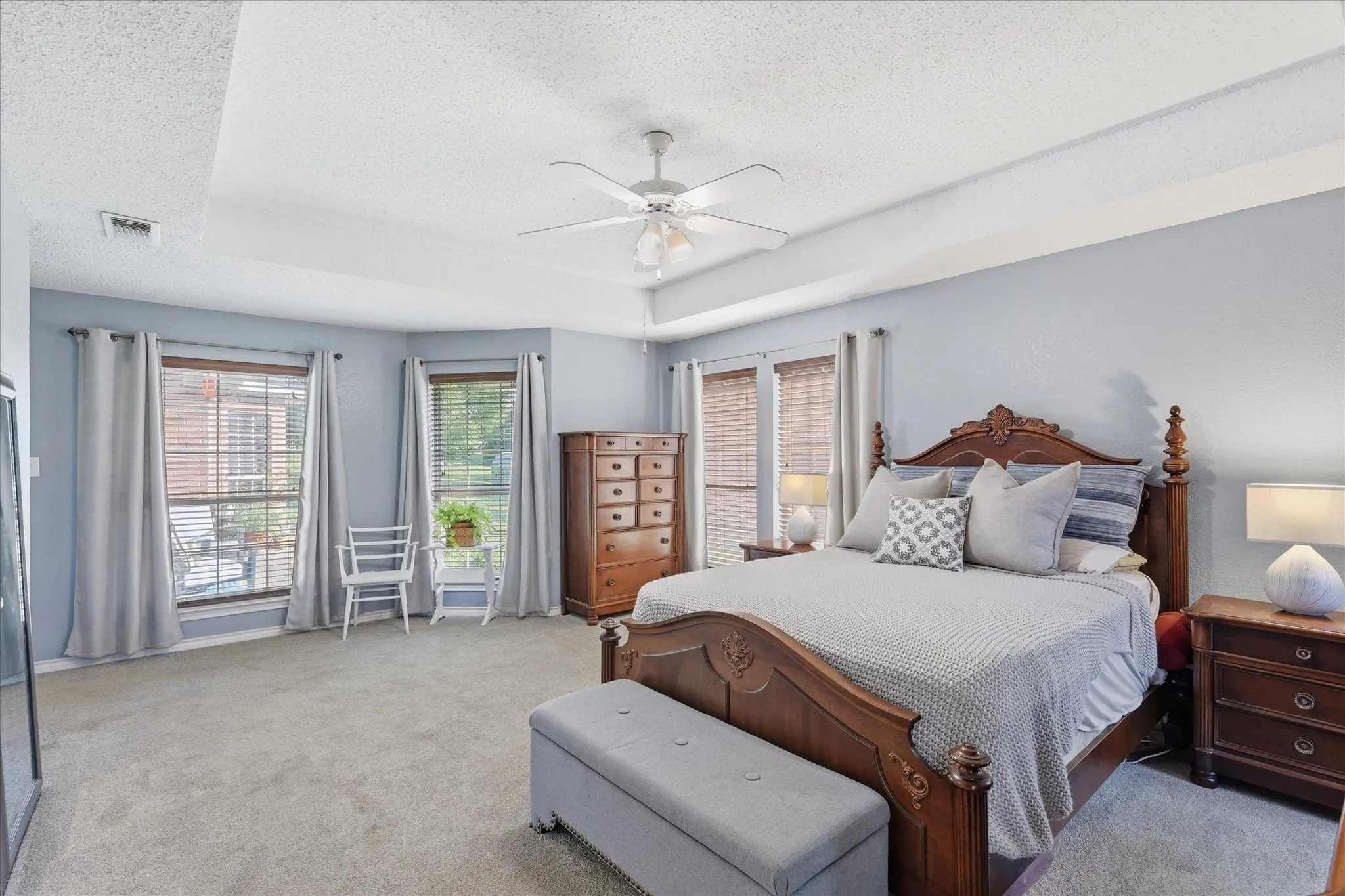 Spacious bedroom featuring a tray ceiling with a ceiling fan, plush carpet flooring, and two large windows with blinds and drapery