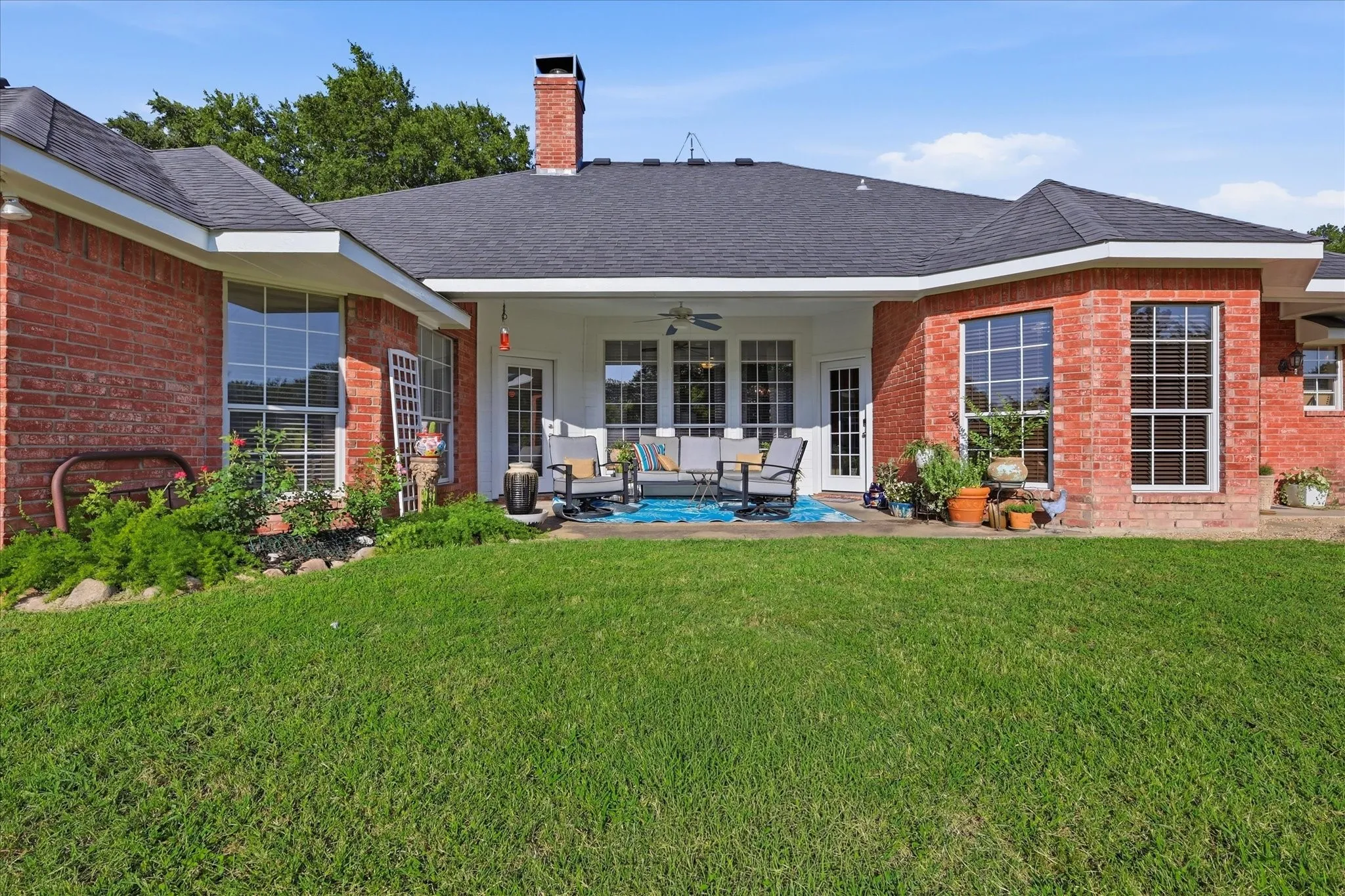 Expansive backyard featuring a manicured lawn, a covered patio, brick exterior, large windows, and mature landscaping