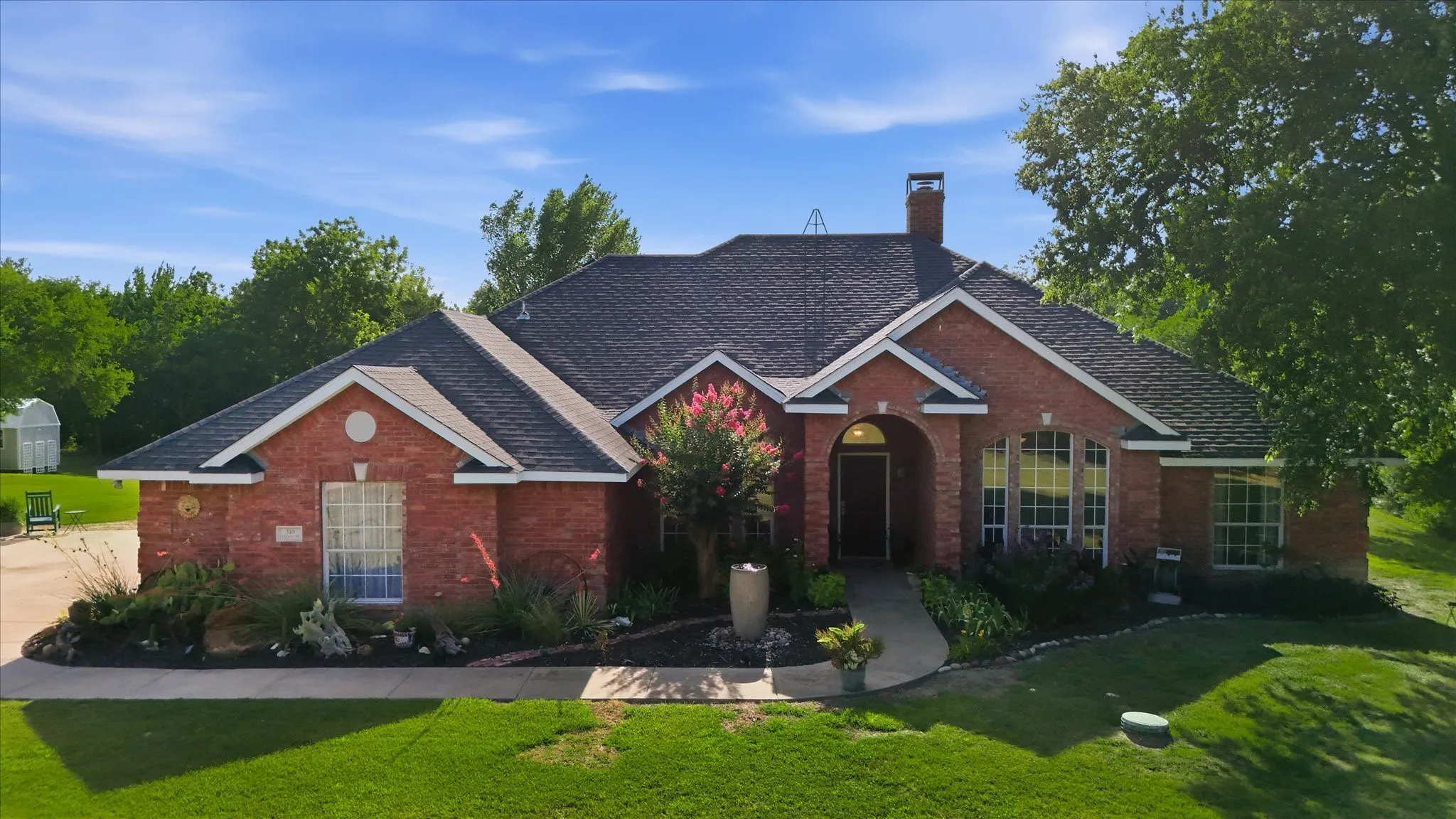 Brick residence featuring a prominent entryway arch, arched windows, a dark shingle roof, and mature landscaping