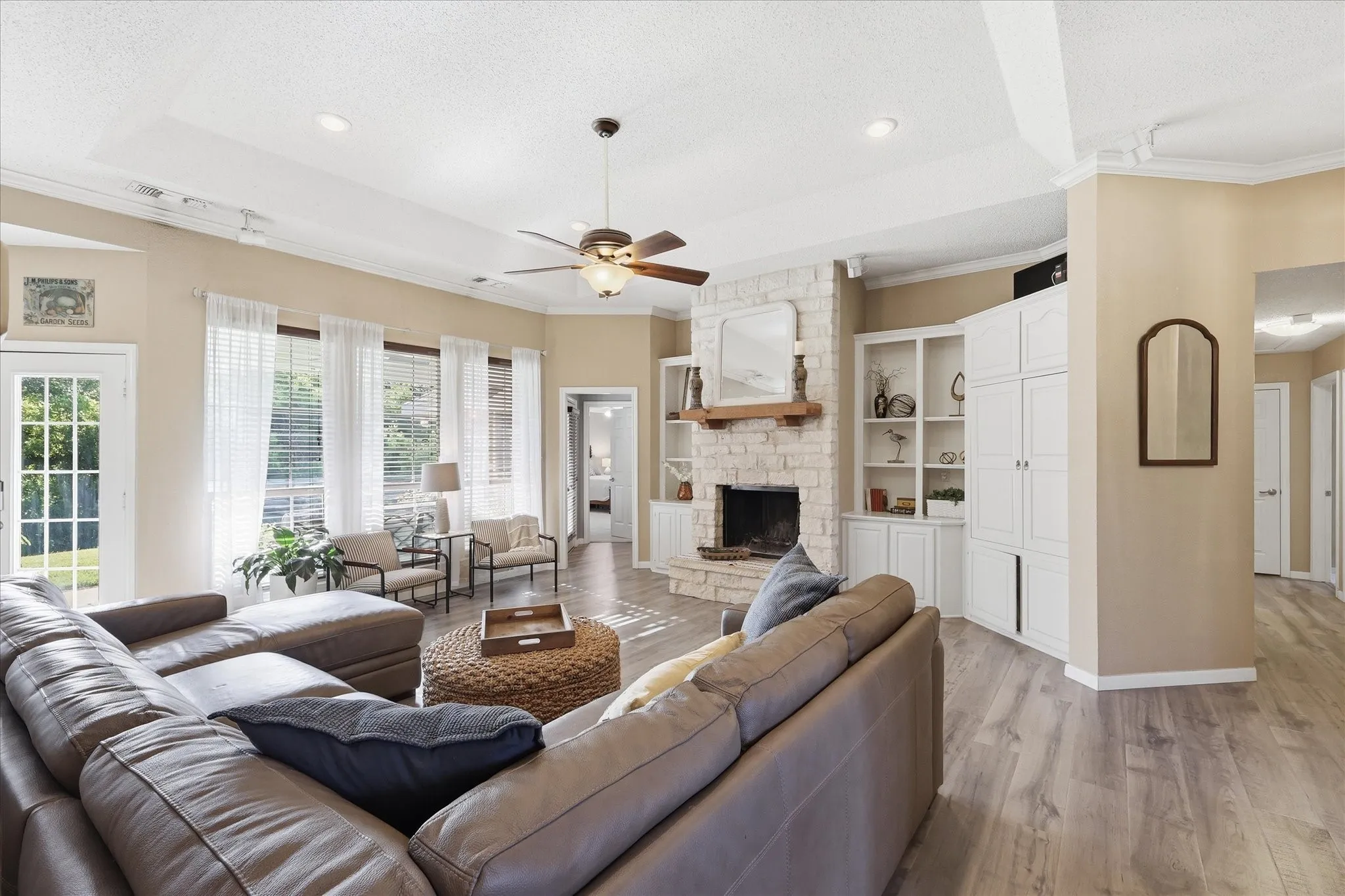 Spacious living area featuring a stone-clad fireplace with wood mantel, built-in cabinetry, wood-finish flooring, a ceiling fan, and multiple windows