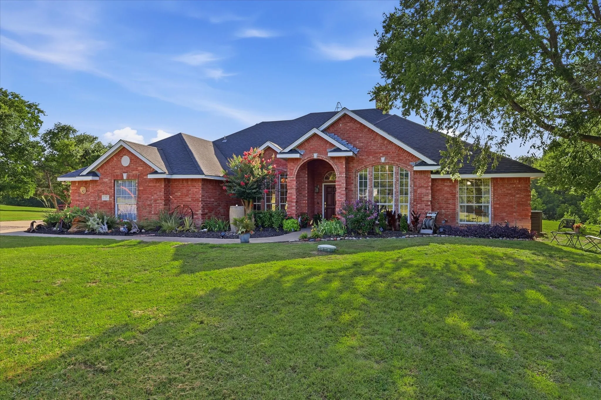 Red brick exterior with a dark shingle roof, arched entry, multiple large windows, and landscaped garden beds