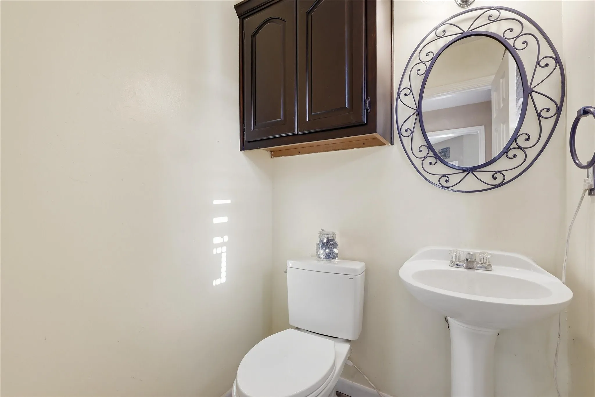 Pedestal sink with dual-handle chrome faucet, oval wall mirror with decorative metal frame, and wall-mounted dark wood cabinetry