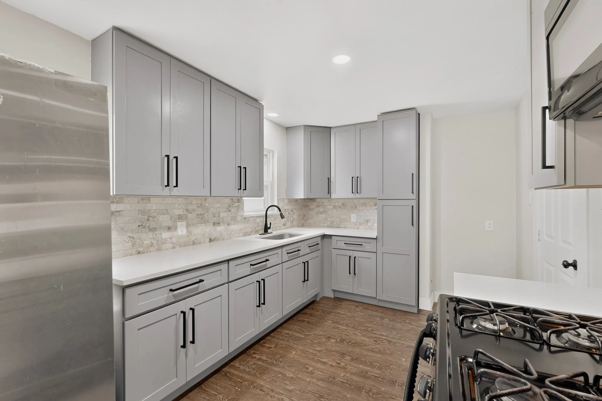 Modern kitchen featuring light gray shaker cabinetry with matte black hardware, white countertops, a mosaic tile backsplash, and wood-finish flooring