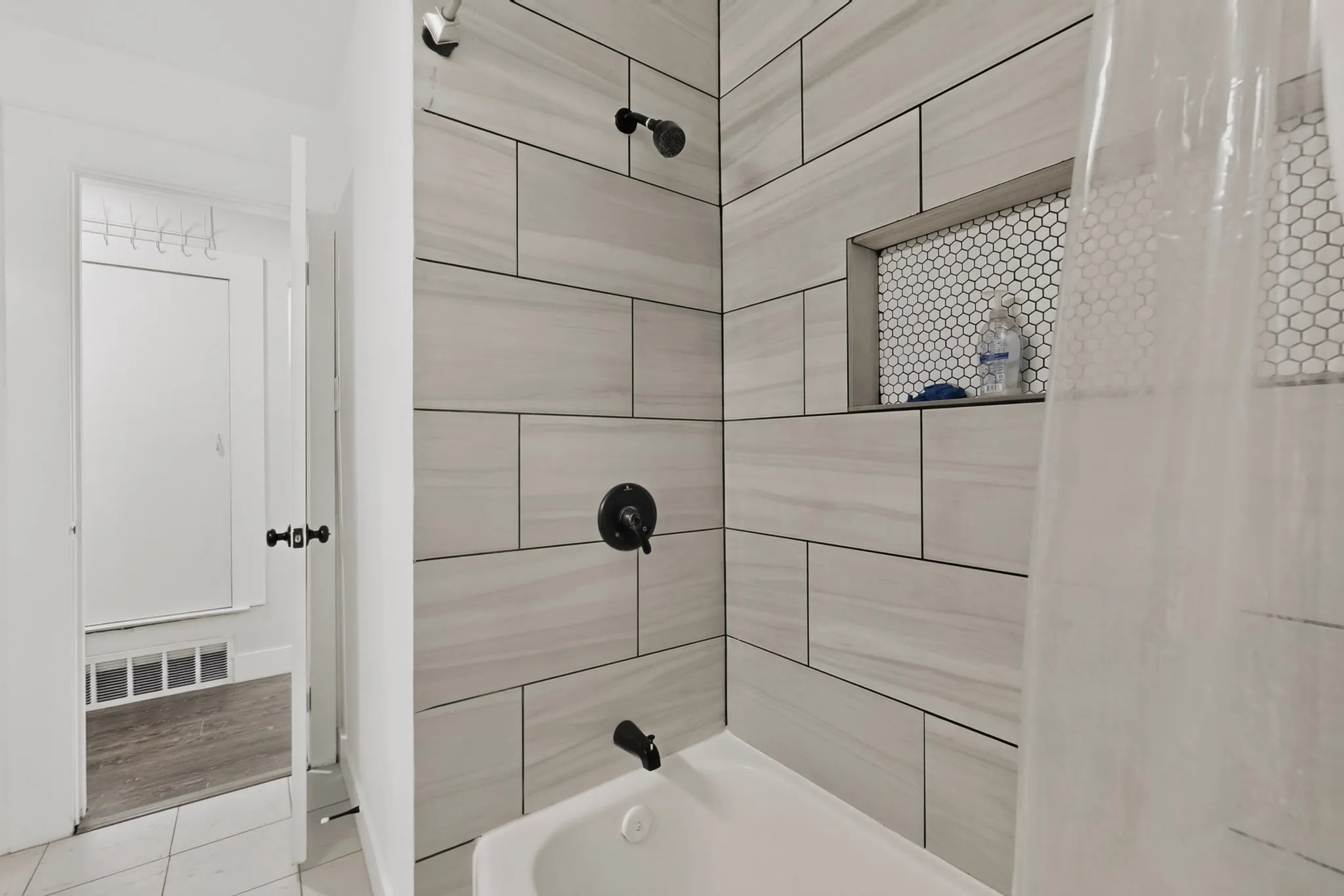 Bathtub with light-toned rectangular wall tiling, matte black shower fixtures, and a tiled shower niche with hexagonal mosaic accents