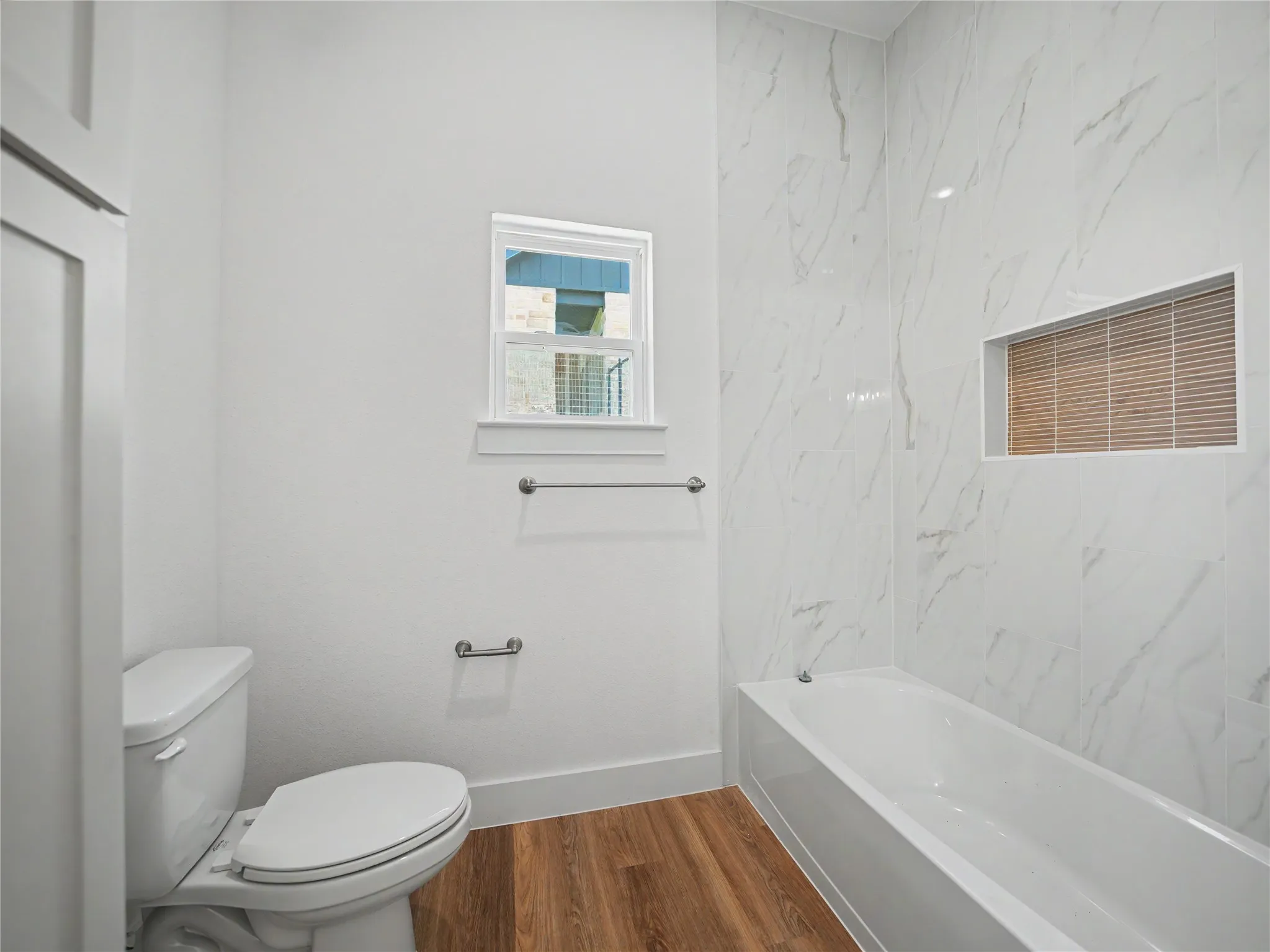 Bathroom featuring a built-in tub, white subway tile surround with a recessed niche, wood-finish flooring, and a window