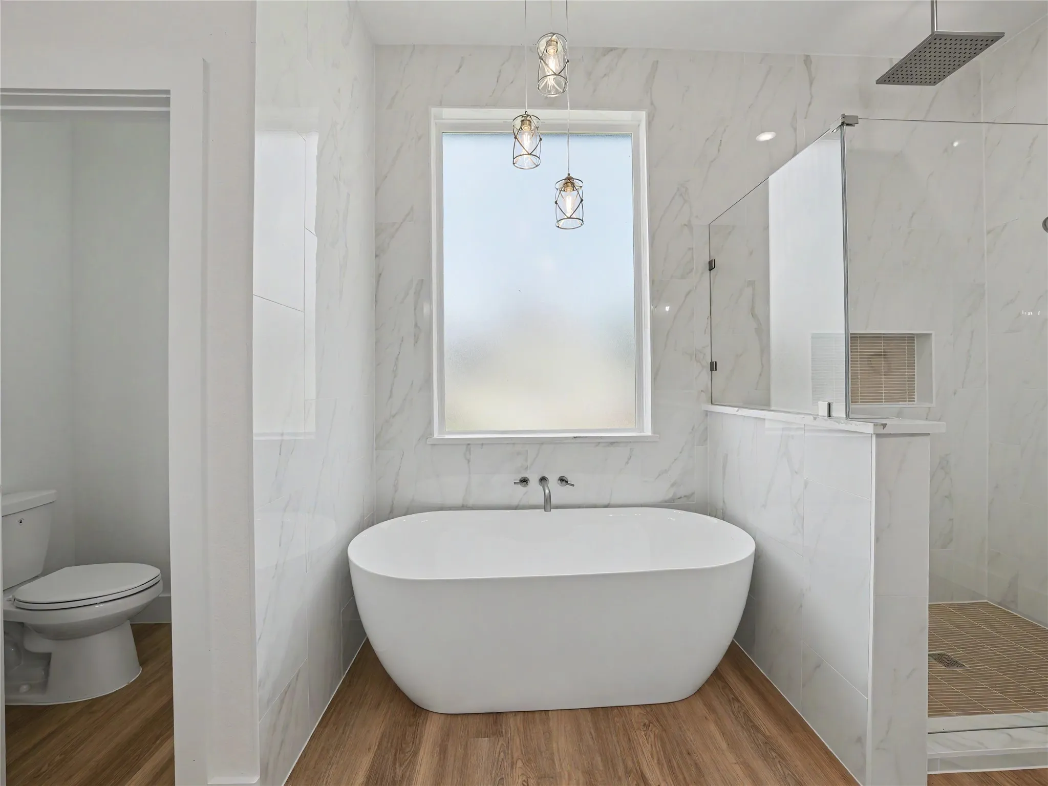 Freestanding soaking tub with wall-mounted faucet, surrounded by marbled-finish tile walls and wood-finish flooring