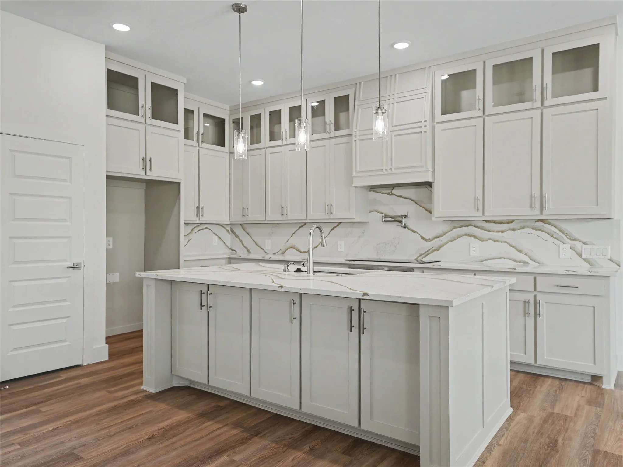 Kitchen featuring light-toned cabinetry, a spacious island with a sink, elegant pendant lighting, a patterned backsplash, and wood-finish flooring