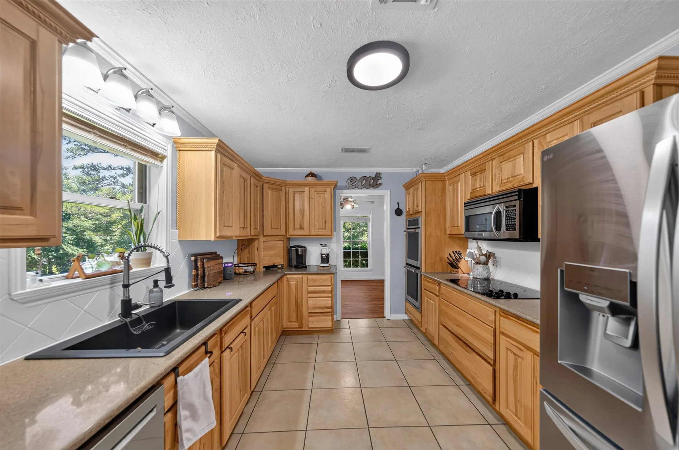 Spacious kitchen featuring extensive light wood cabinetry, light-colored tile flooring, a large window, a black composite sink with a gooseneck faucet, and stainless steel appliances including a double oven