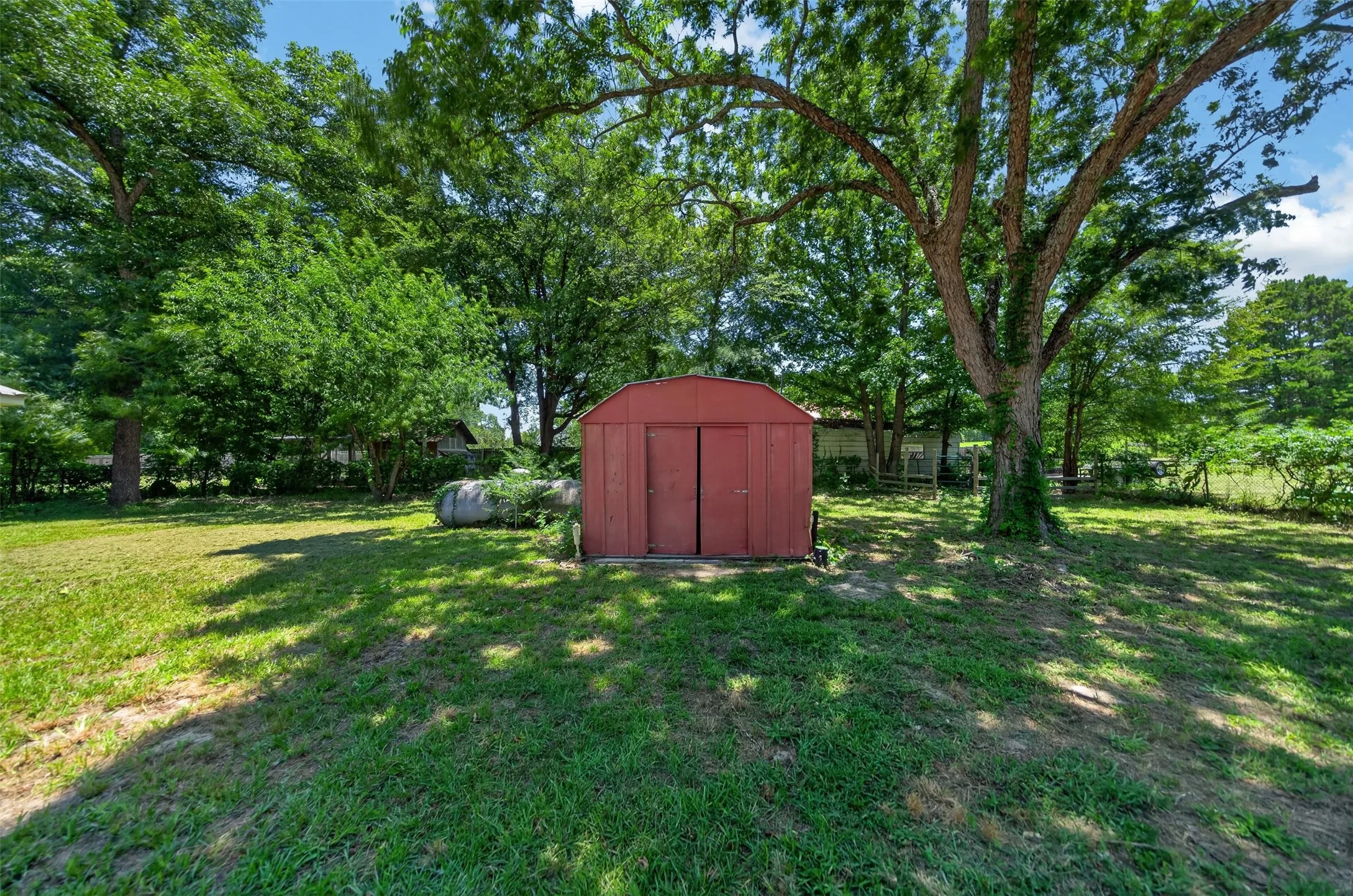 Expansive lawn area featuring a red storage shed with double doors, mature trees providing natural shade, and a visible fence line