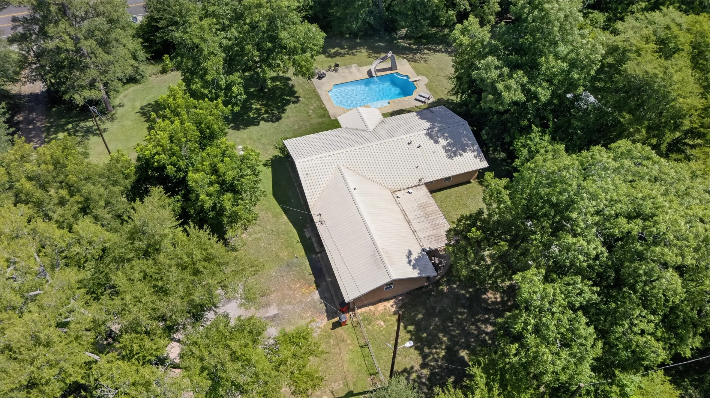 Spacious property featuring a light-colored metal roof, private swimming pool, surrounding green lawn, and mature trees