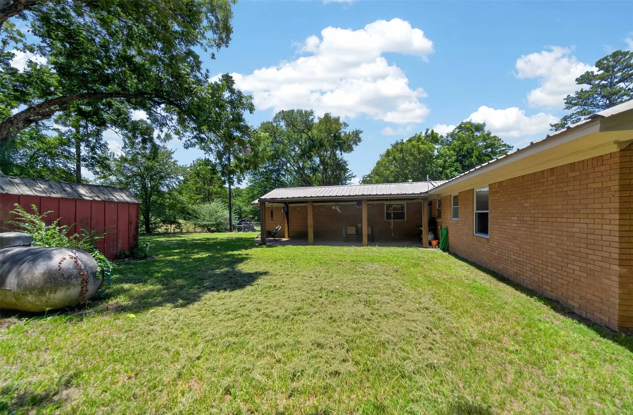 Spacious backyard featuring a covered patio, a red storage shed, and mature trees