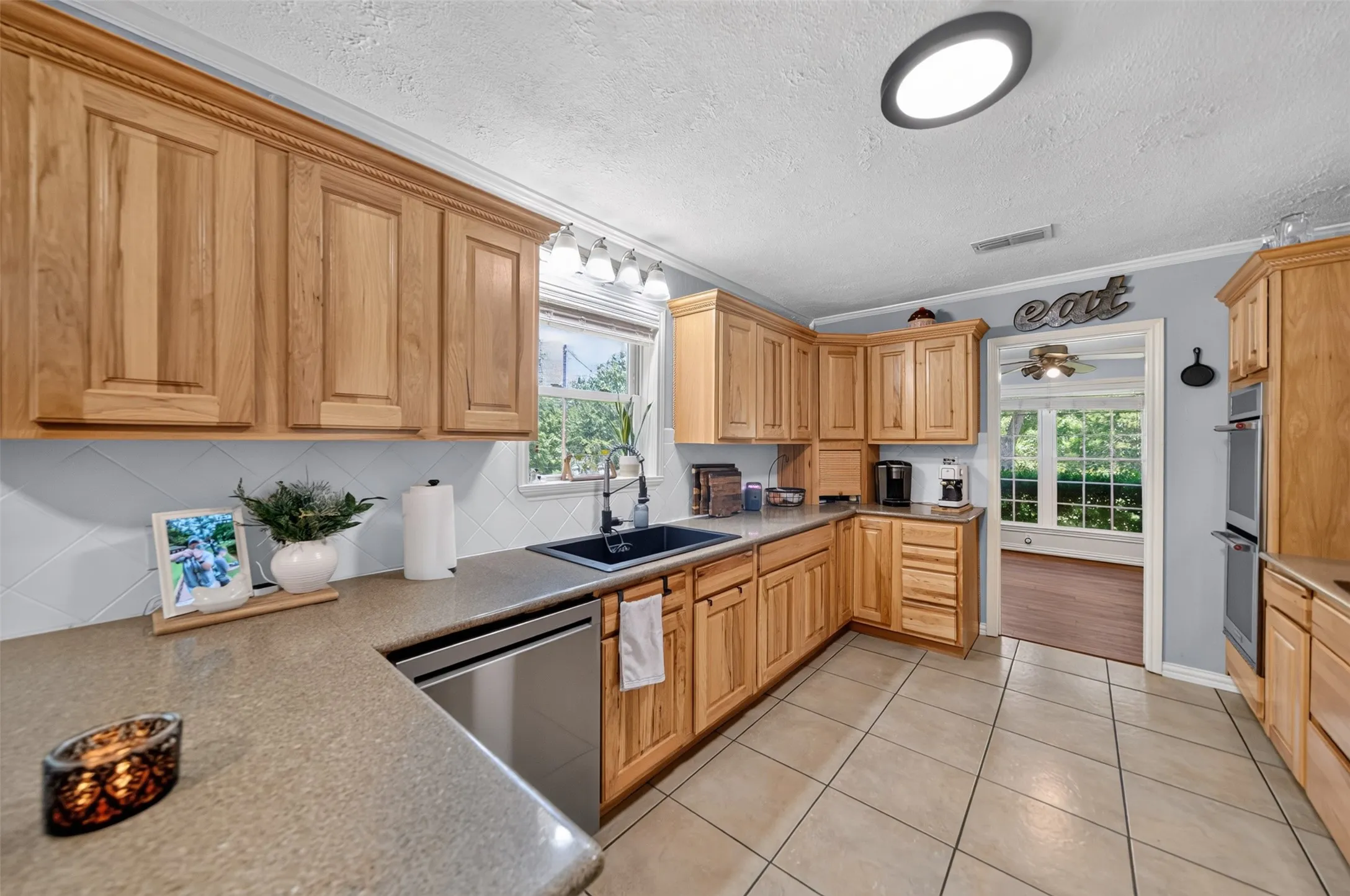 Well-appointed kitchen featuring extensive light wood cabinetry, stone-look countertops, a stainless steel dishwasher, and a double wall oven